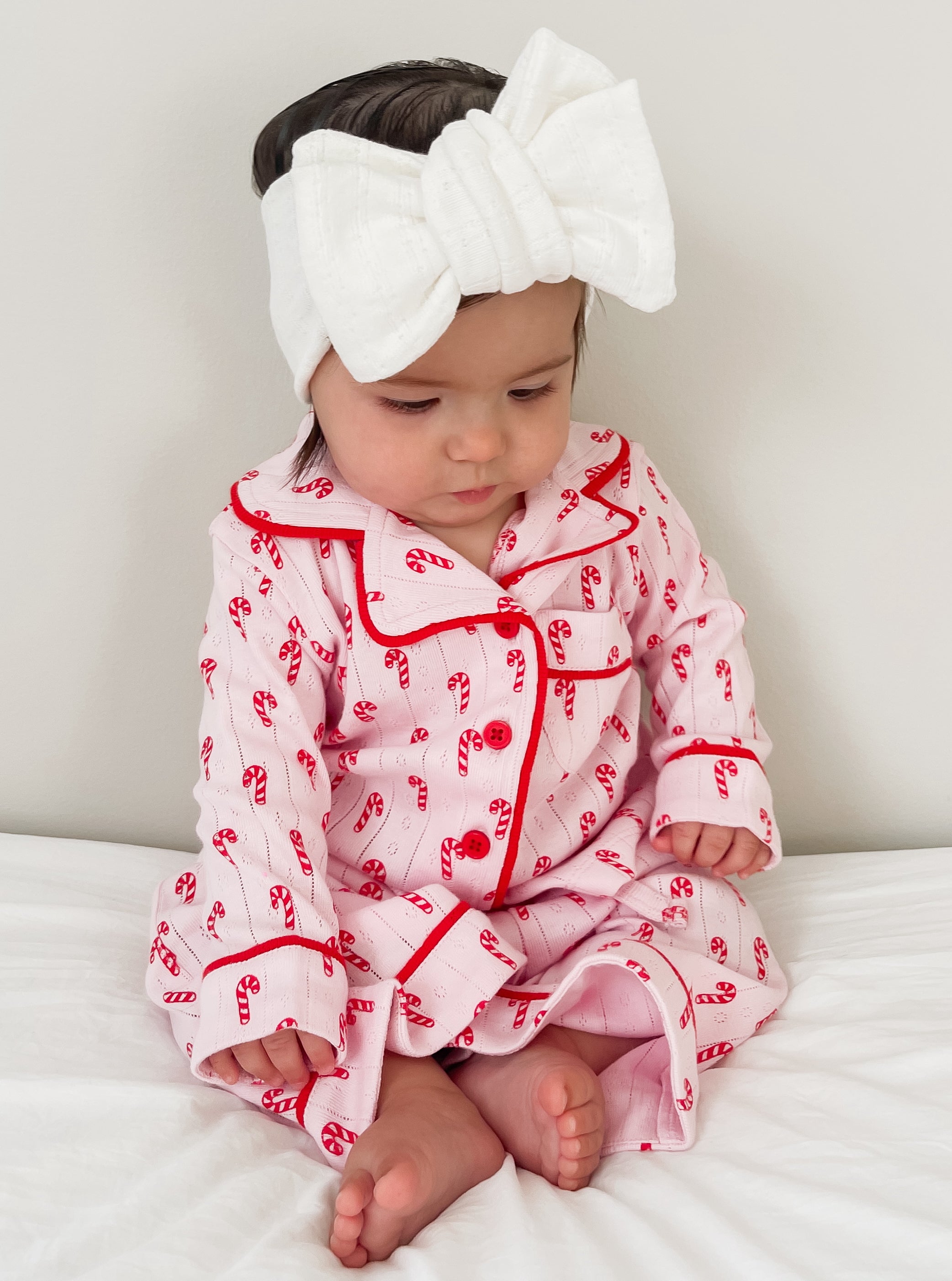 Baby girl in pink candy cane pajamas and a white bow headband sits on a white bed, looking down.