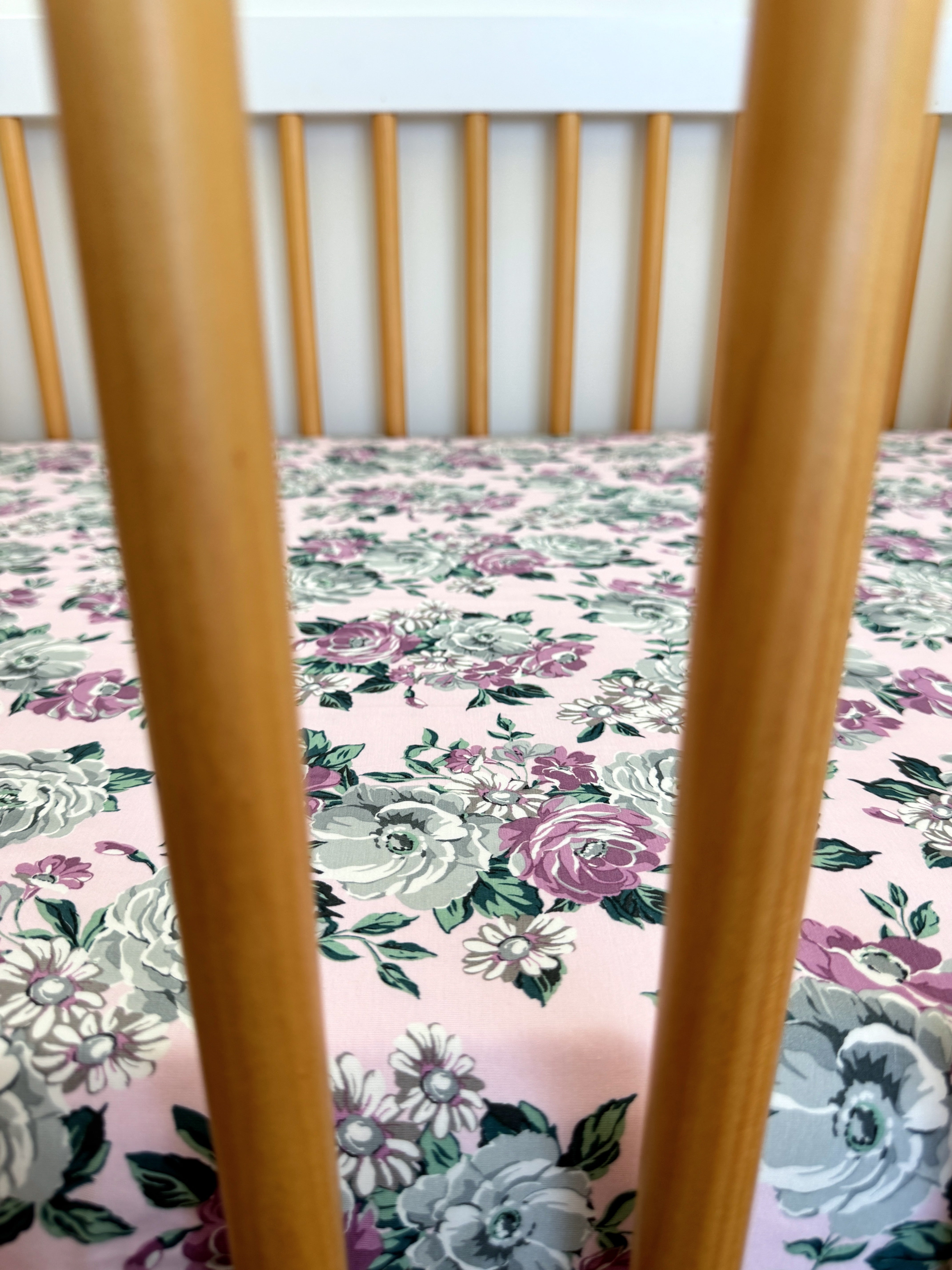 Wooden crib with vertical slats, featuring a floral-patterned pink mattress with gray and purple flowers.
