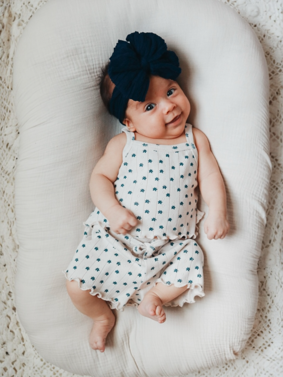 Smiling baby on a cozy surface, wearing a floral outfit and a large dark bow headband.