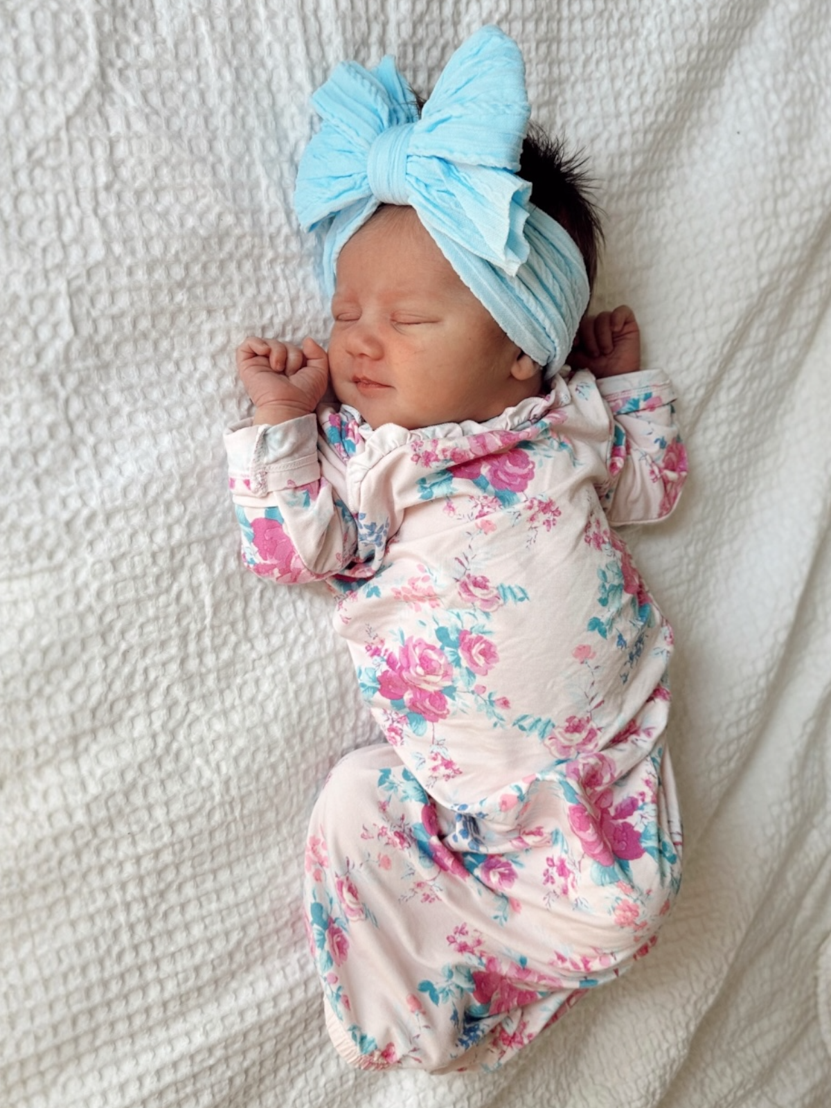 Newborn baby girl in floral pajamas and a large blue bow headband, peacefully sleeping on a white blanket.