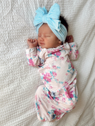 Newborn baby girl in floral pajamas and a large blue bow headband, peacefully sleeping on a white blanket.