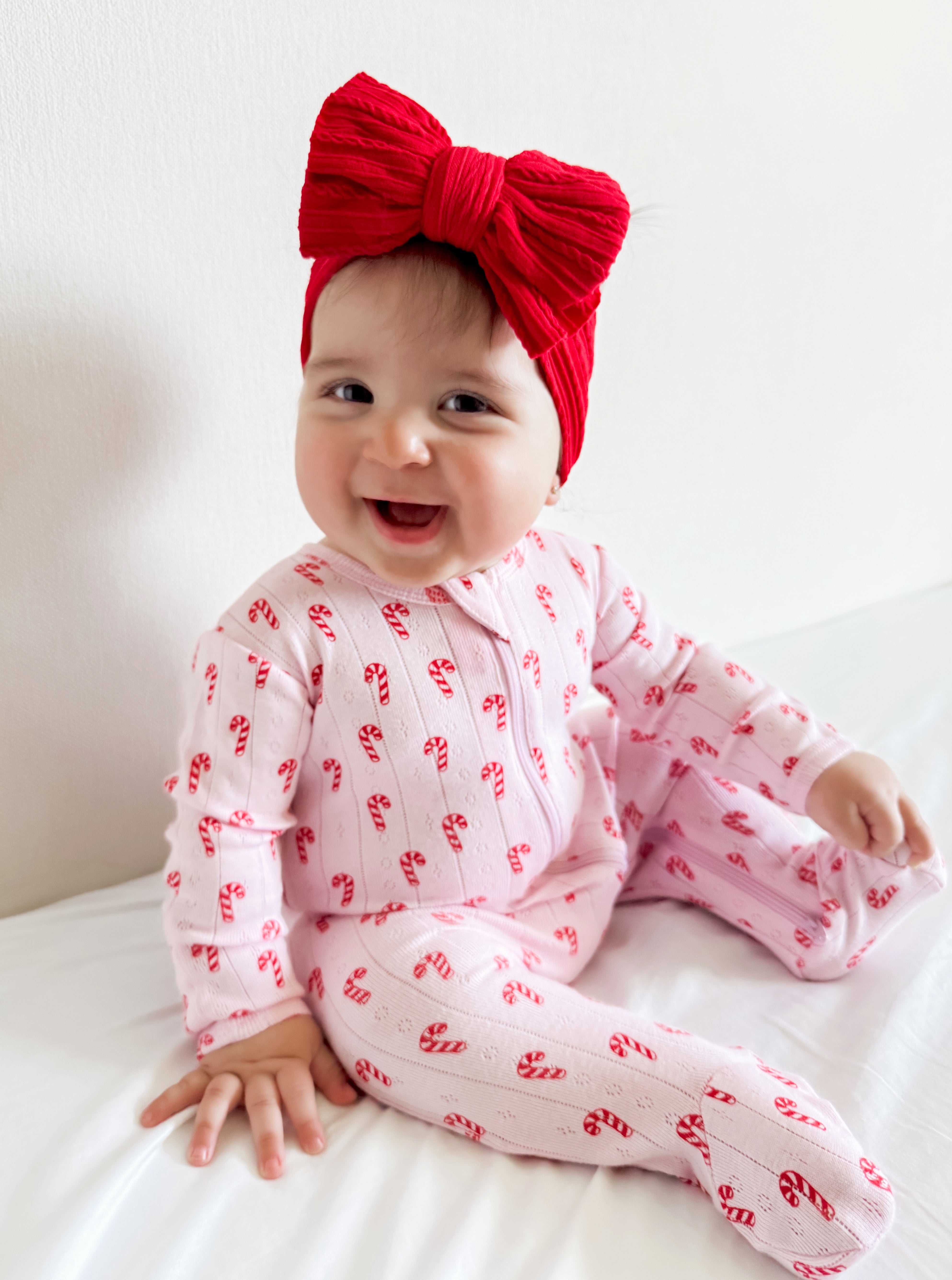 Smiling baby in a pink candy-cane outfit with a large red bow, sitting on a white bed.