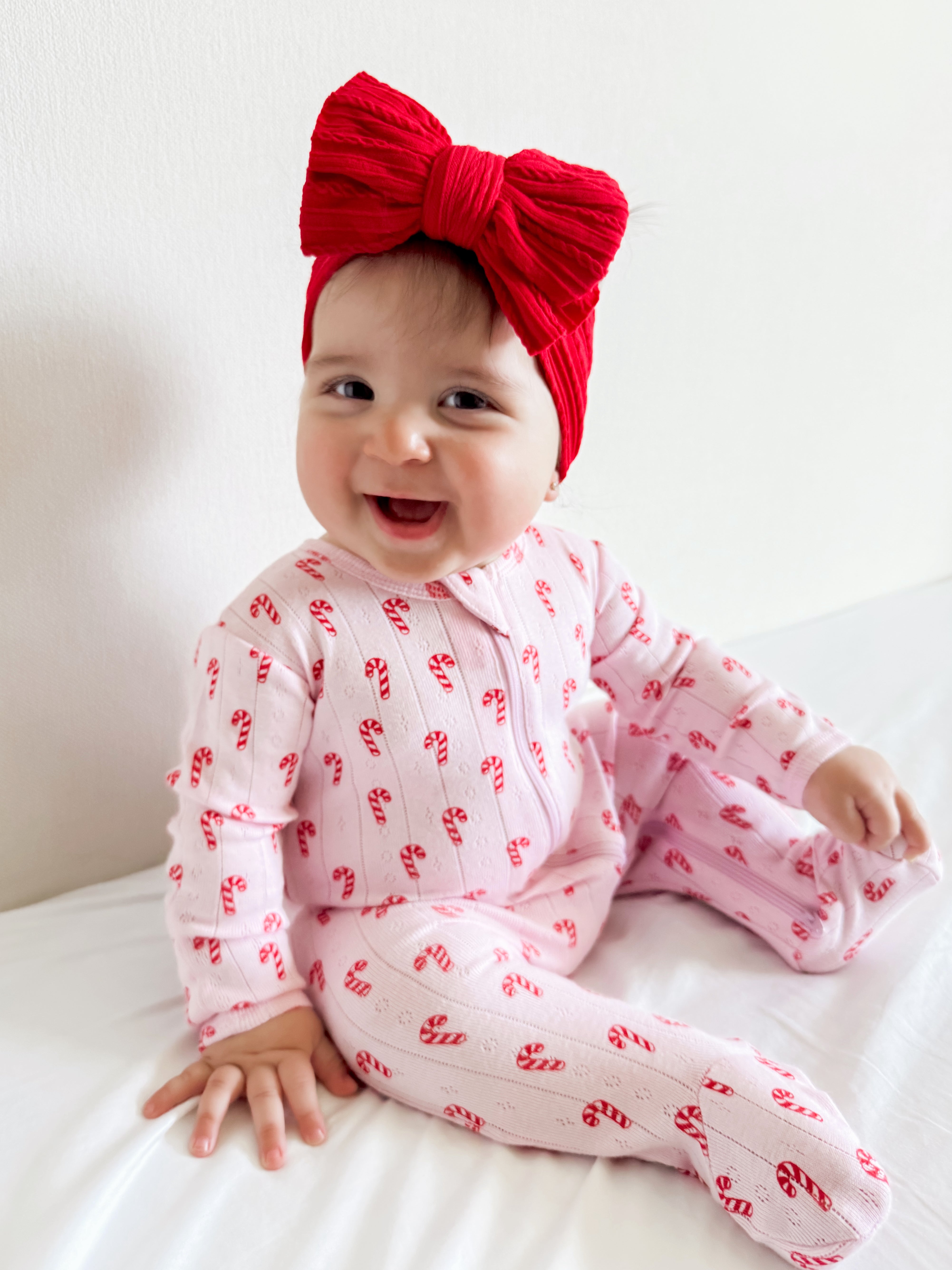 Smiling baby in a pink candy-cane outfit with a large red bow, sitting on a white bed.