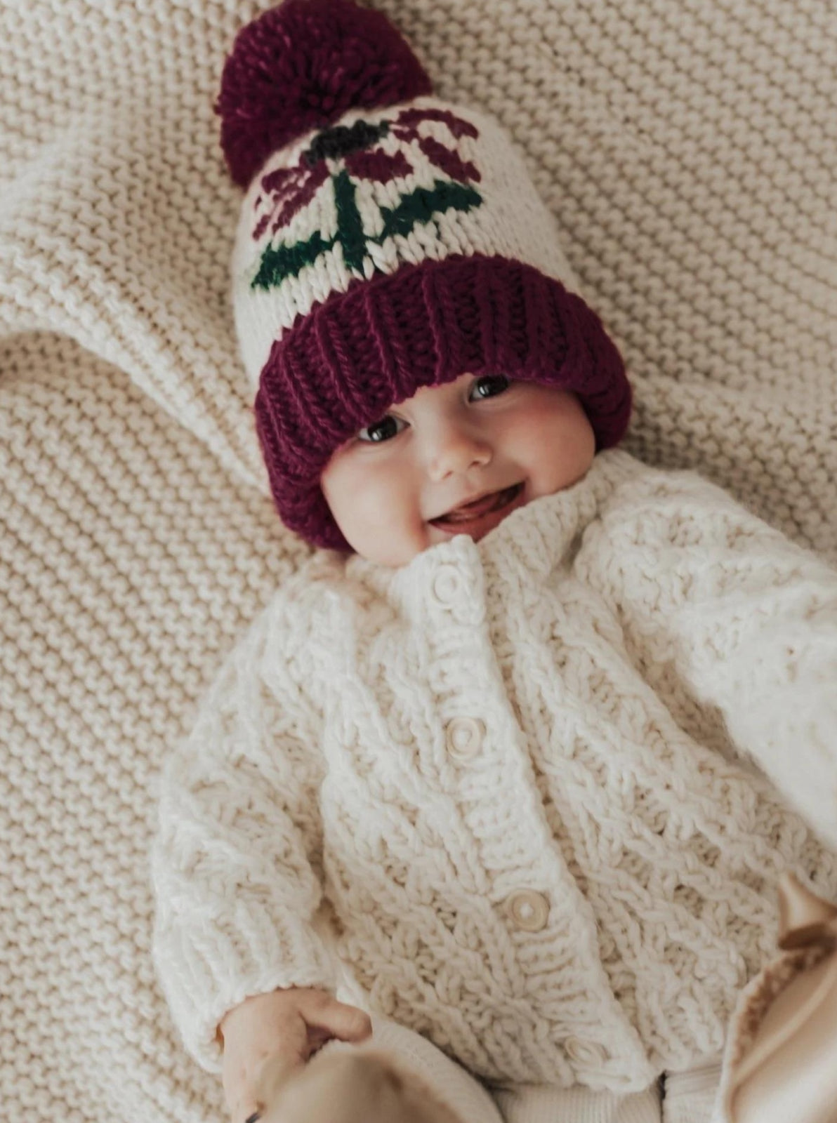 Smiling baby in a knitted cream sweater and colorful beanie on a cozy knitted blanket.