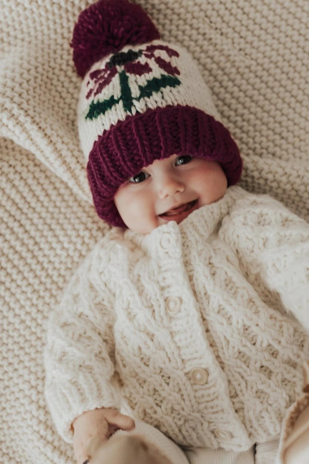 Smiling baby in a knitted cream sweater and colorful beanie on a cozy knitted blanket.
