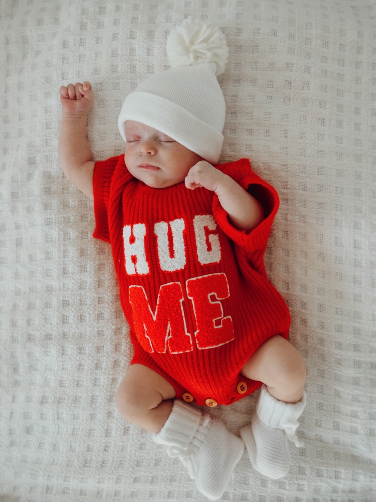 Baby wearing a red "HUG ME" outfit, sleeping peacefully on a textured white blanket.