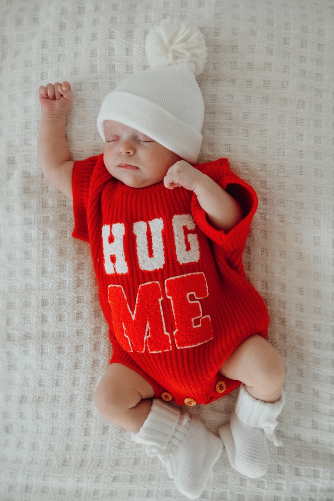 Baby wearing a red "HUG ME" outfit, sleeping peacefully on a textured white blanket.