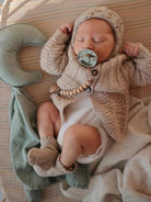 Sleeping baby in a cozy knit outfit with a pacifier, surrounded by soft blankets and a crescent-shaped pillow.