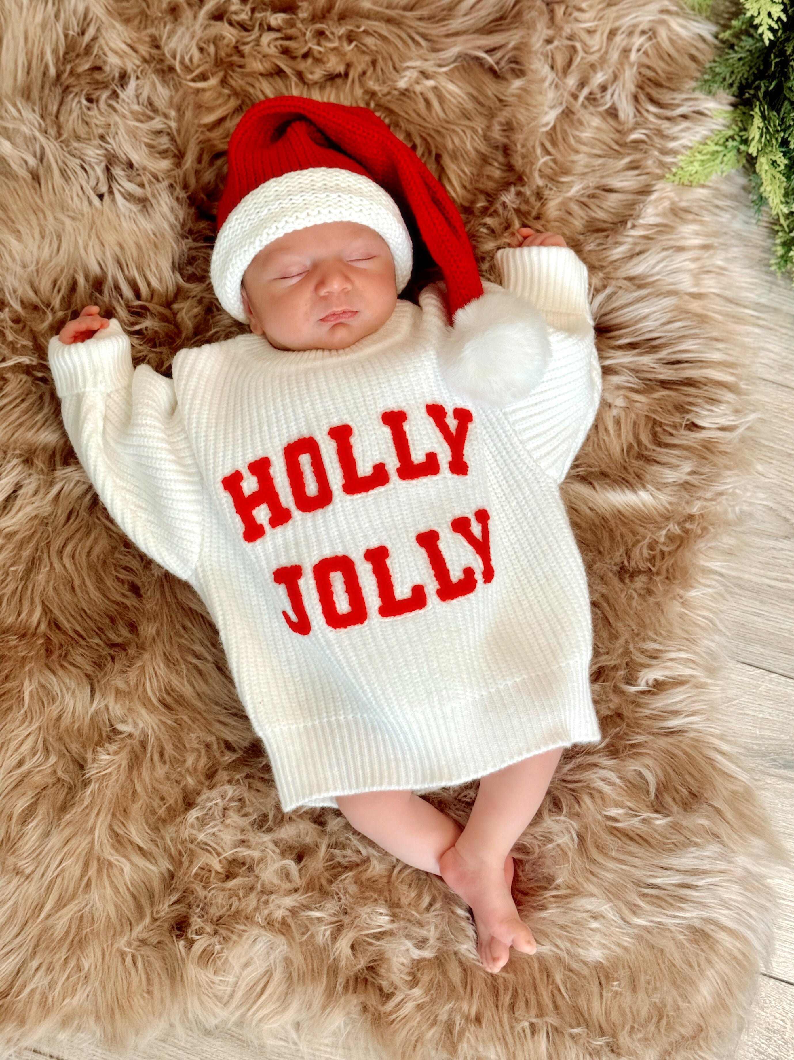 Newborn in a cozy white sweater with "HOLLY JOLLY" text, wearing a red Santa hat, resting on a fluffy rug.