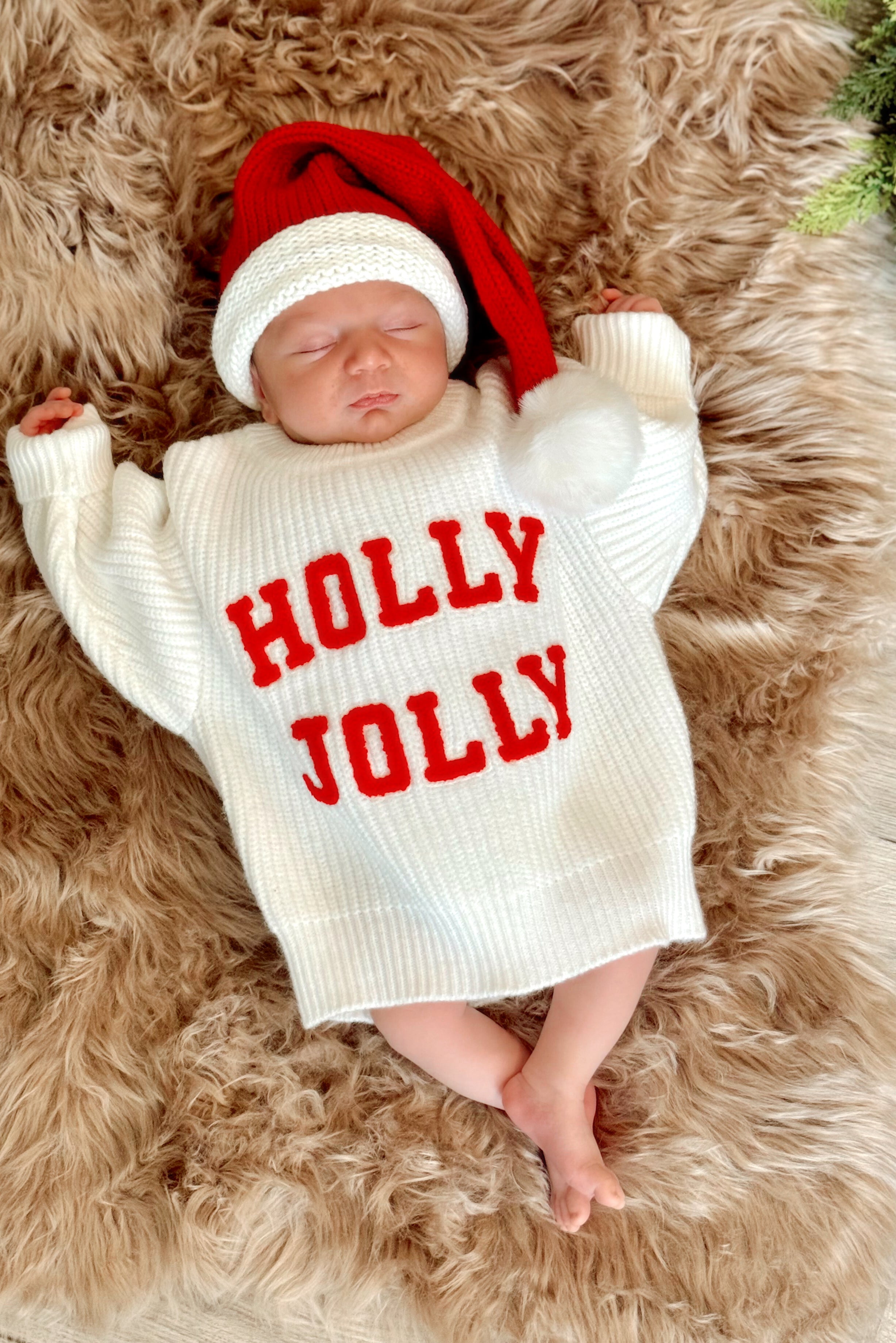 Newborn in a cozy white sweater with "HOLLY JOLLY" text, wearing a red Santa hat, resting on a fluffy rug.