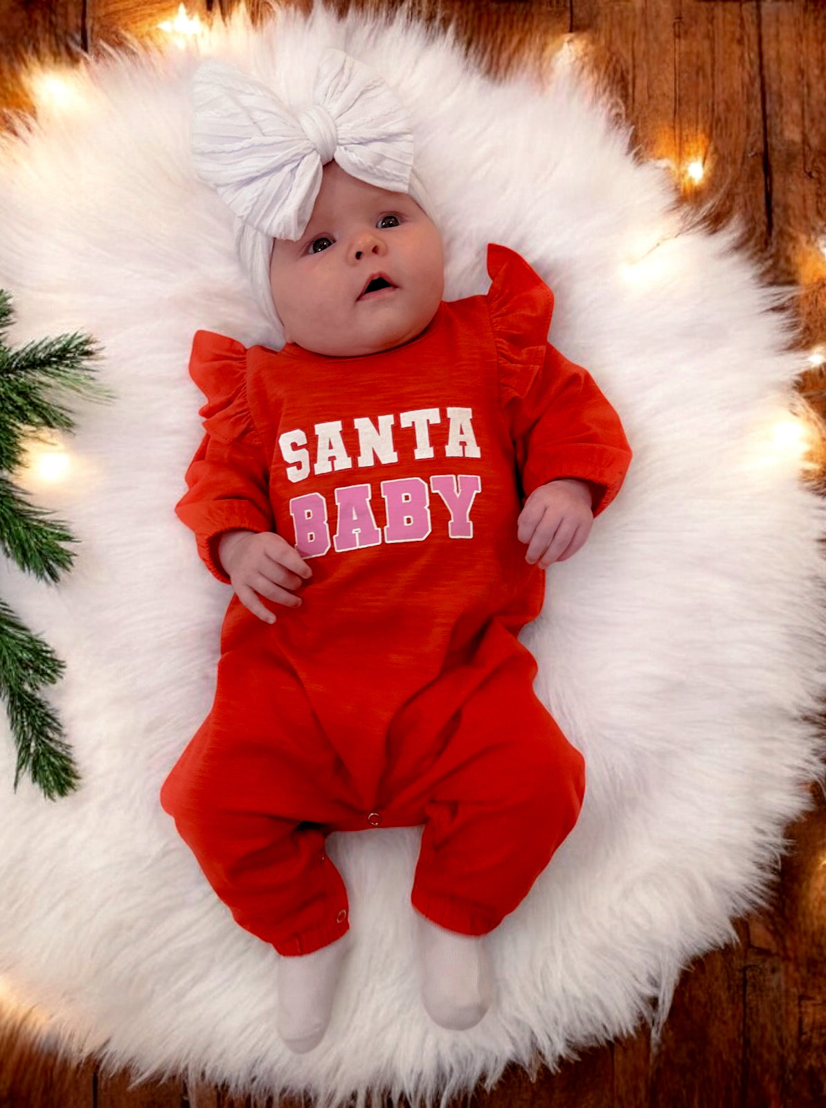 Baby wearing a red "Santa Baby" outfit with a large white bow, resting on a fluffy white rug with holiday lights.