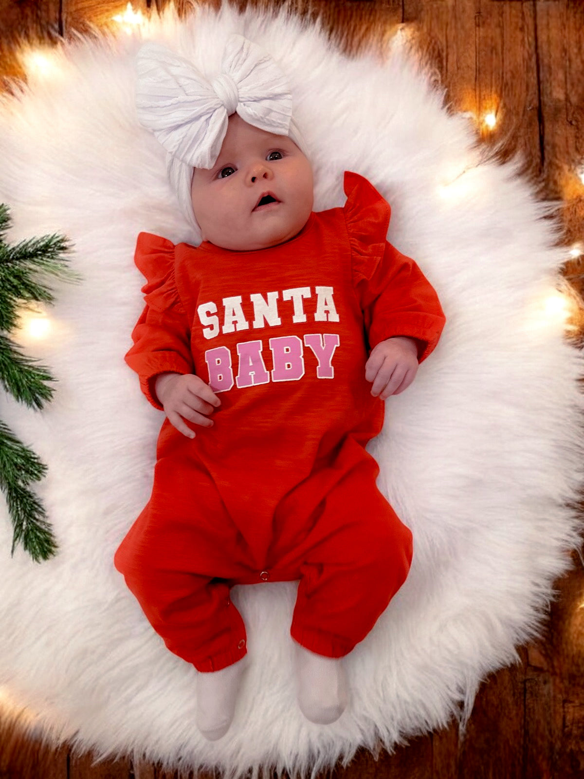 Baby wearing a red "Santa Baby" outfit with a large white bow, resting on a fluffy white rug with holiday lights.