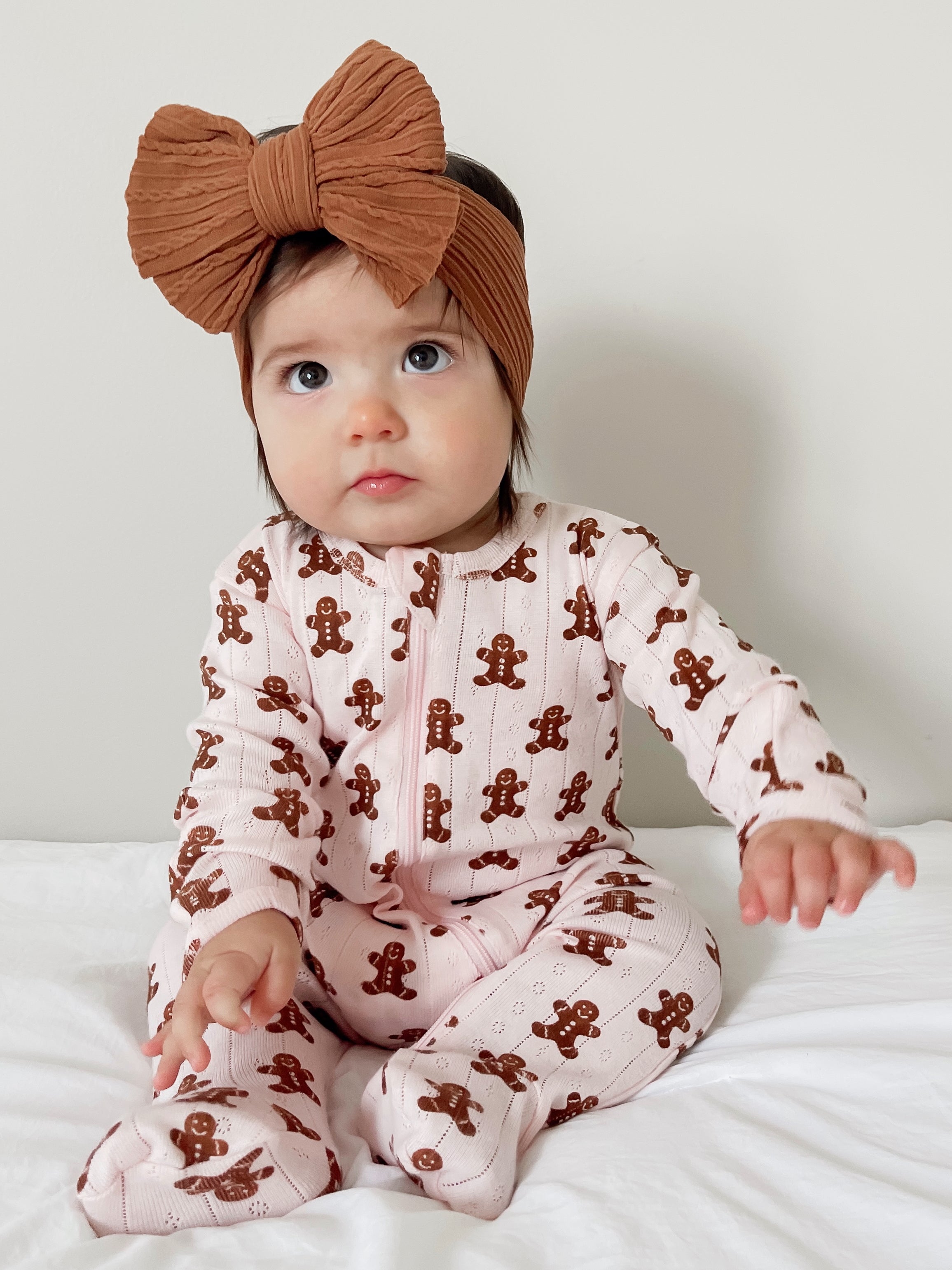 Baby girl in pink gingerbread-patterned pajamas and a brown headband, sitting on a white blanket.