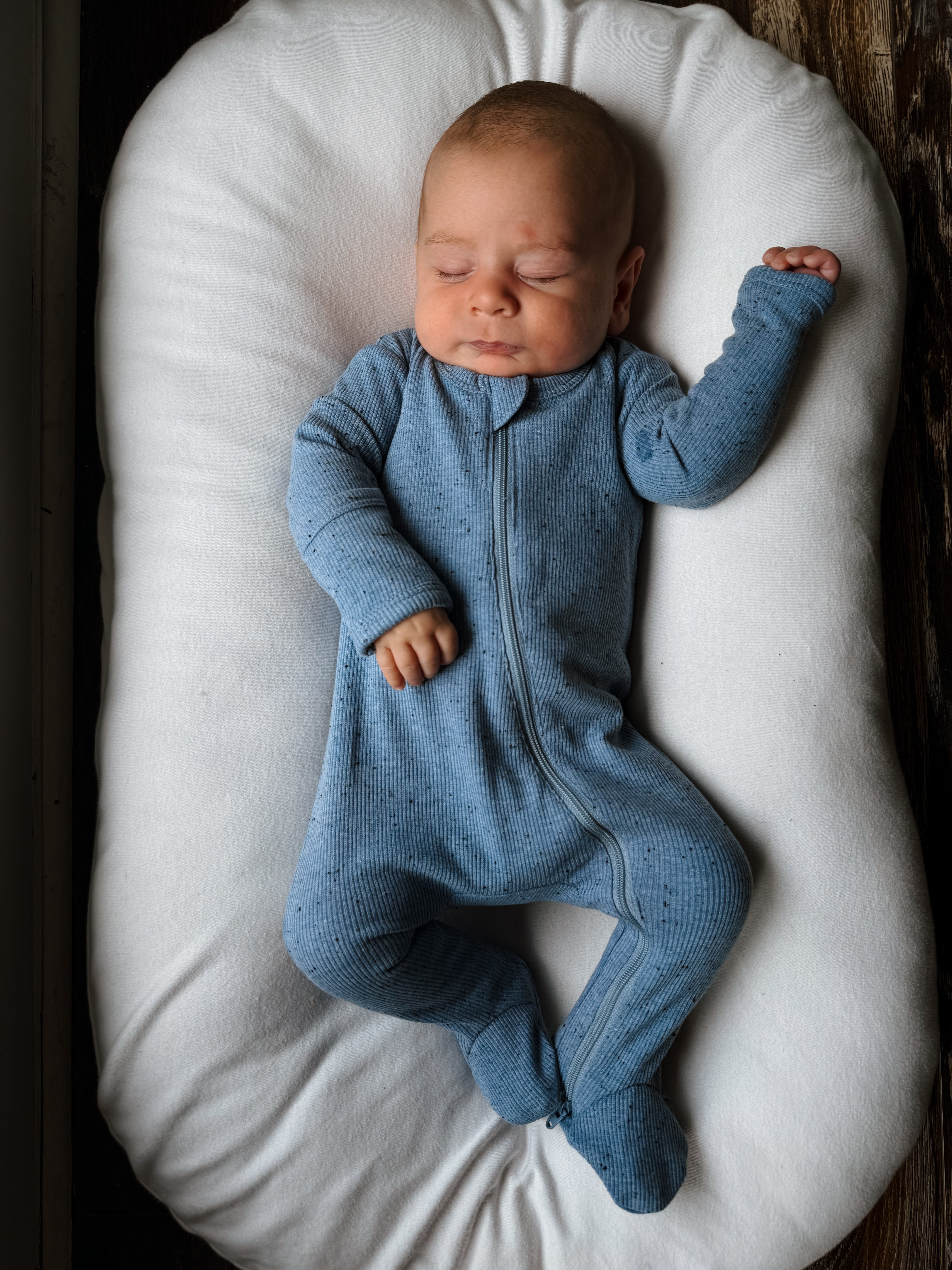 A sleeping baby in a blue onesie lies on a white pillow on a wooden surface.