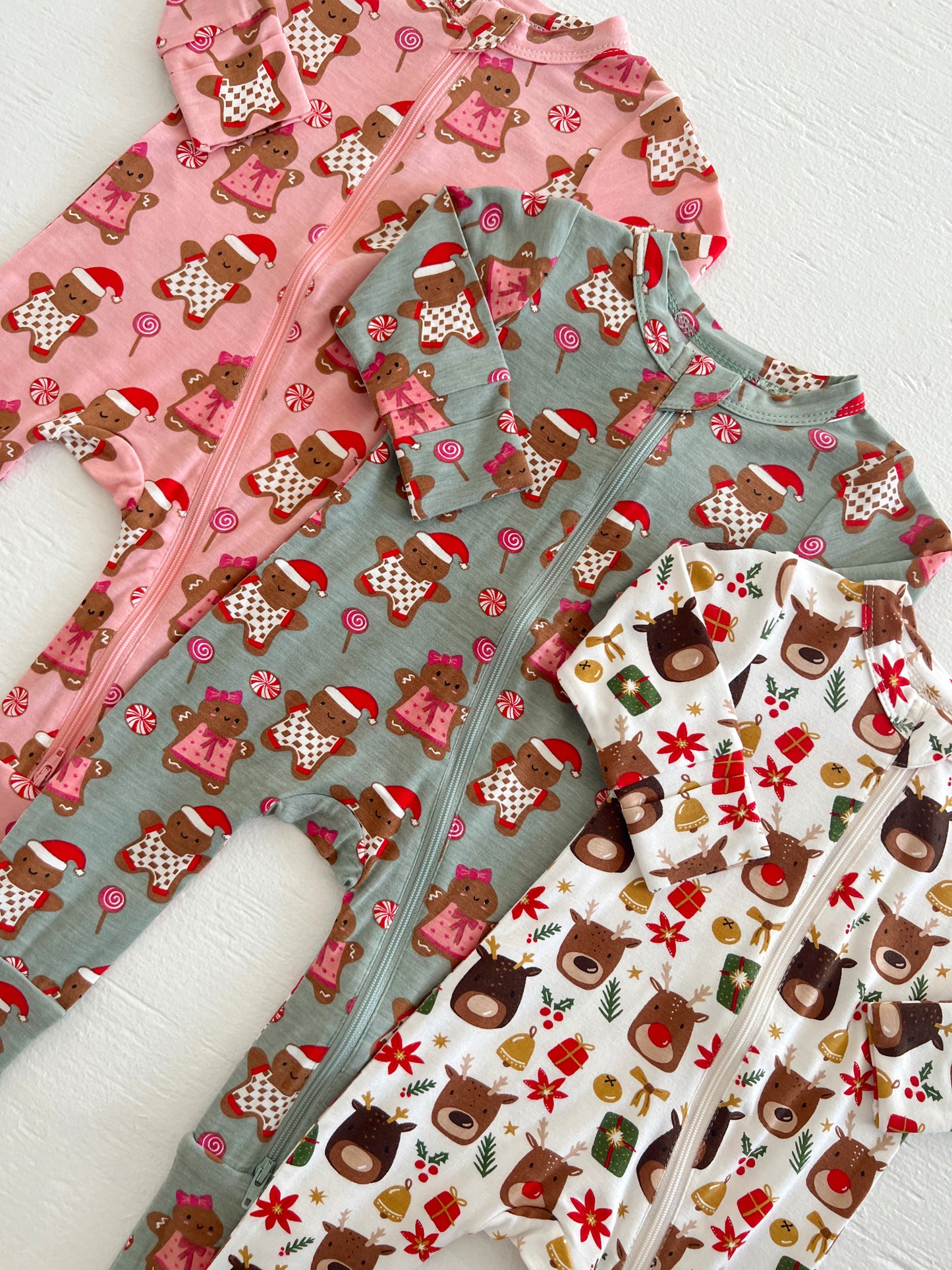 Three festive children's onesies featuring gingerbread men, reindeer, and holiday designs in pink, green, and white.