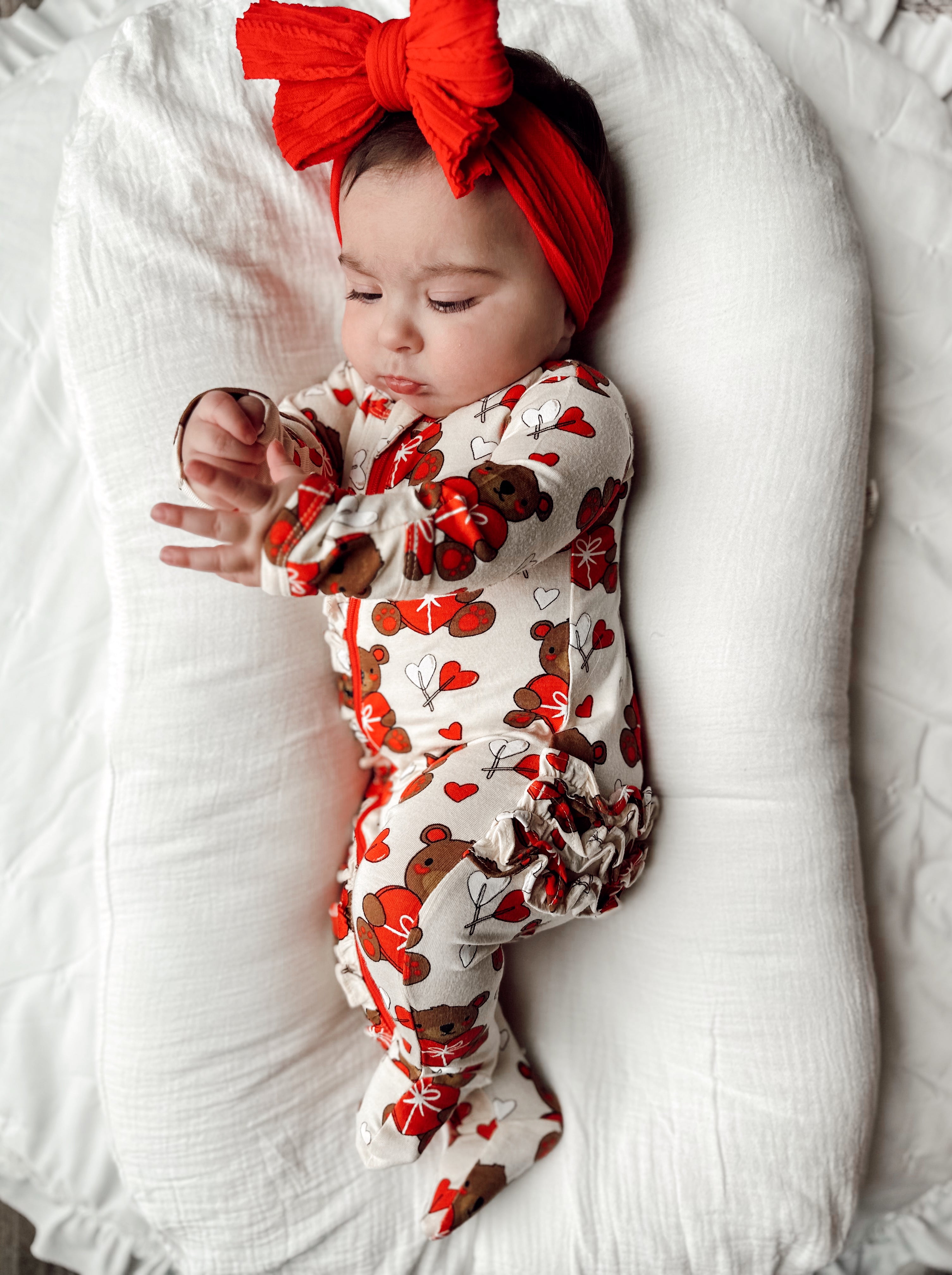 Baby in a floral onesie with a red bow, resting on a white surface.