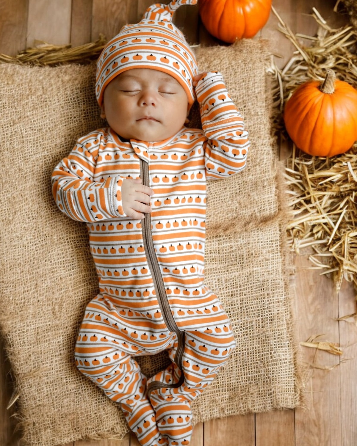 Sleeping baby in pumpkin-patterned outfit, surrounded by pumpkins on a straw mat. Warm autumn theme.