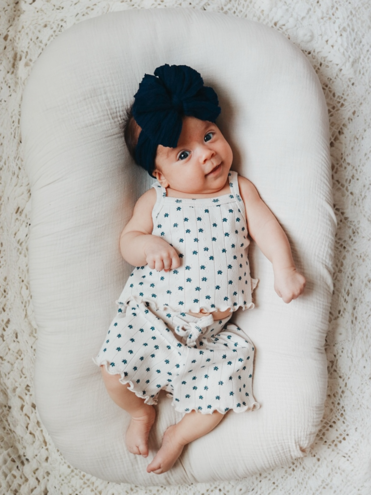 Smiling baby in a patterned outfit and headband, resting on a cozy textured backdrop.