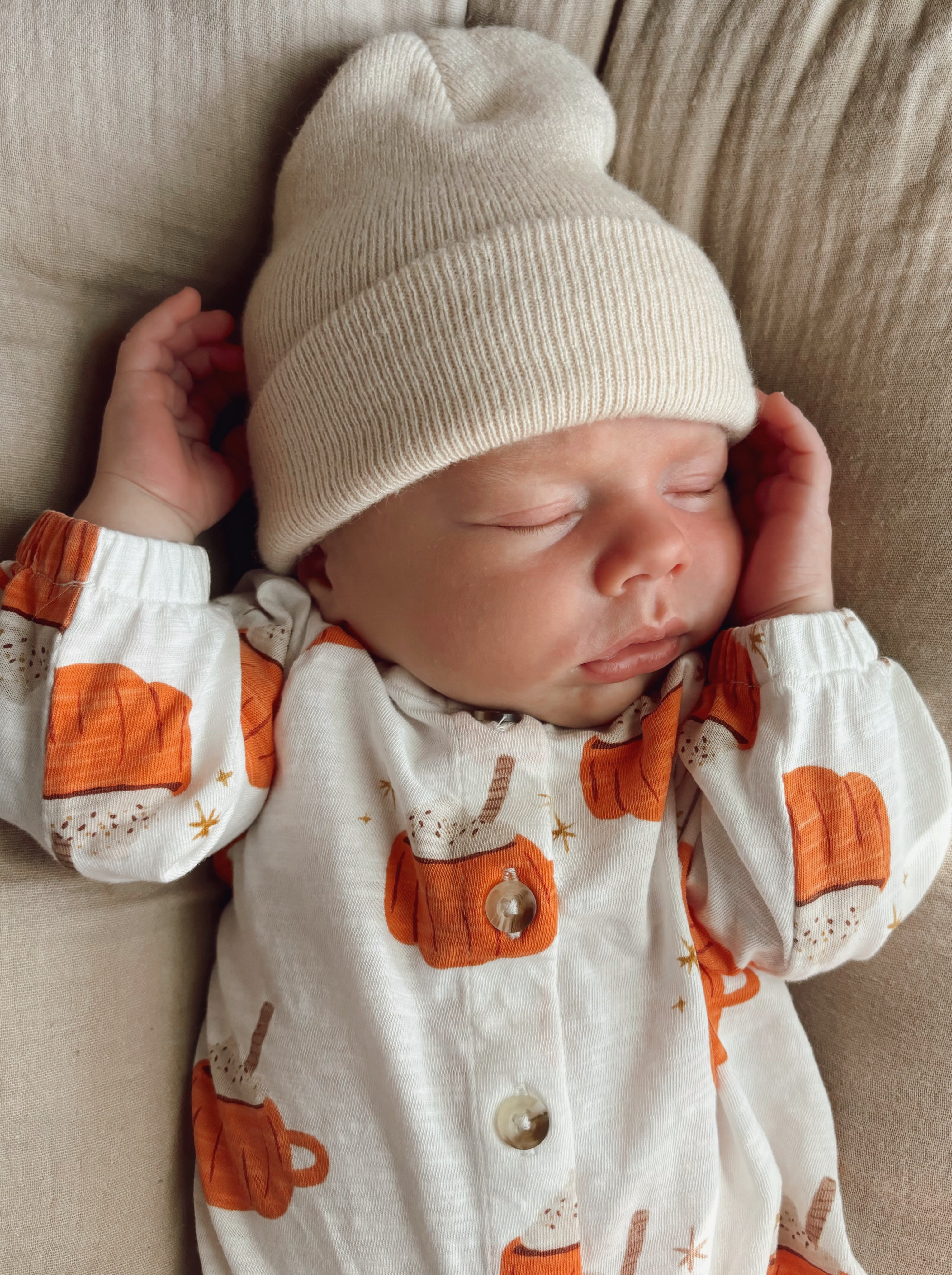 Sleeping baby in a pumpkin-patterned outfit and cream beanie, resting peacefully on a beige surface.