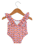 Pink swimsuit with orange fruit pattern, featuring ruffled straps, hanging on a wooden hanger.