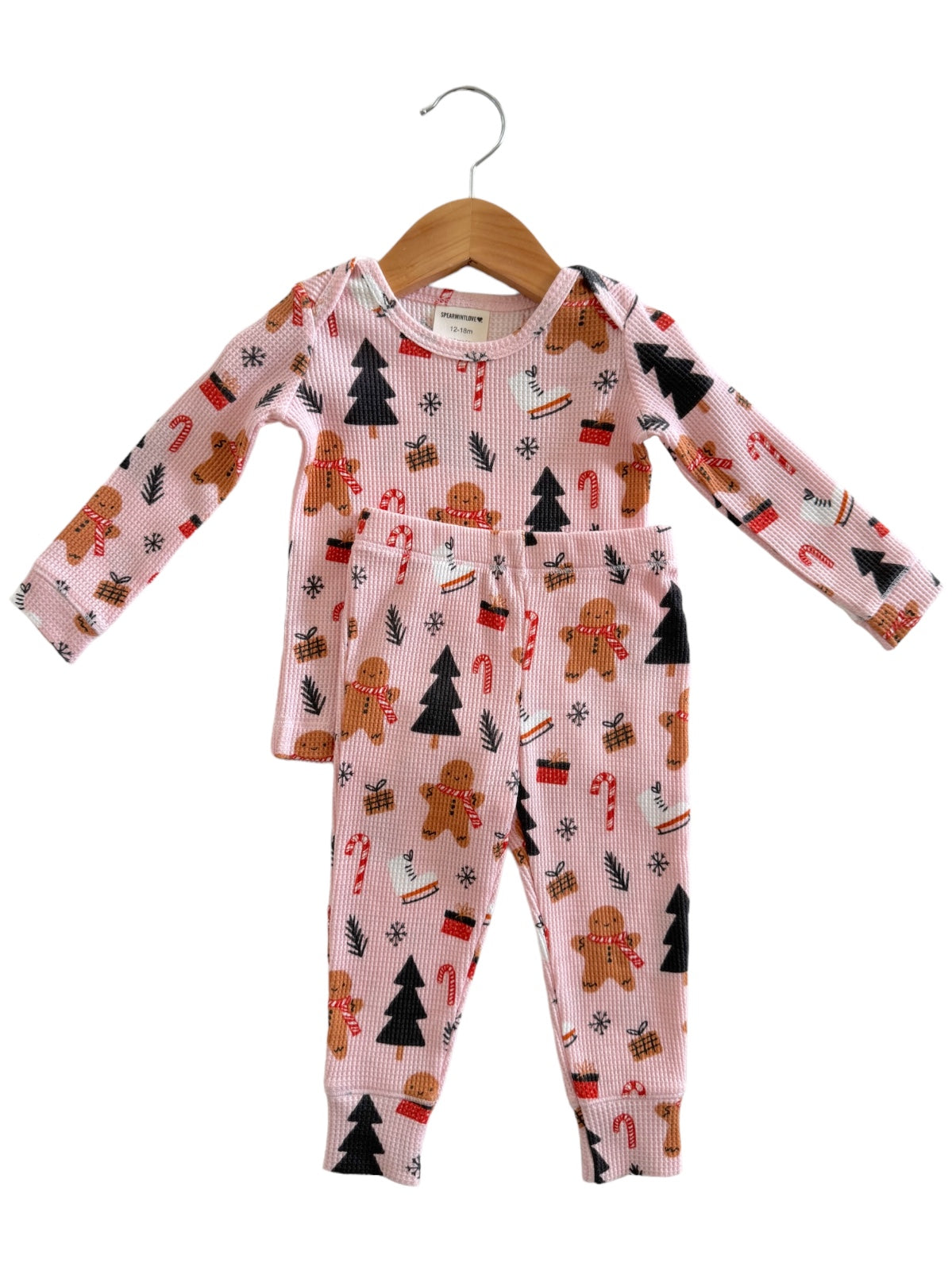 Pink pajama set featuring gingerbread, candy canes, and Christmas trees, hanging on a wooden hanger.