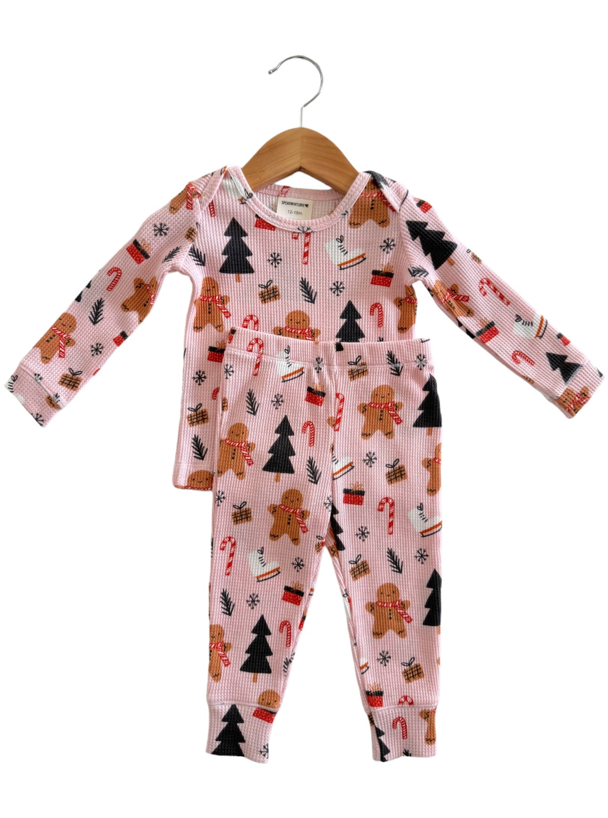 Pink pajama set featuring gingerbread, candy canes, and Christmas trees, hanging on a wooden hanger.