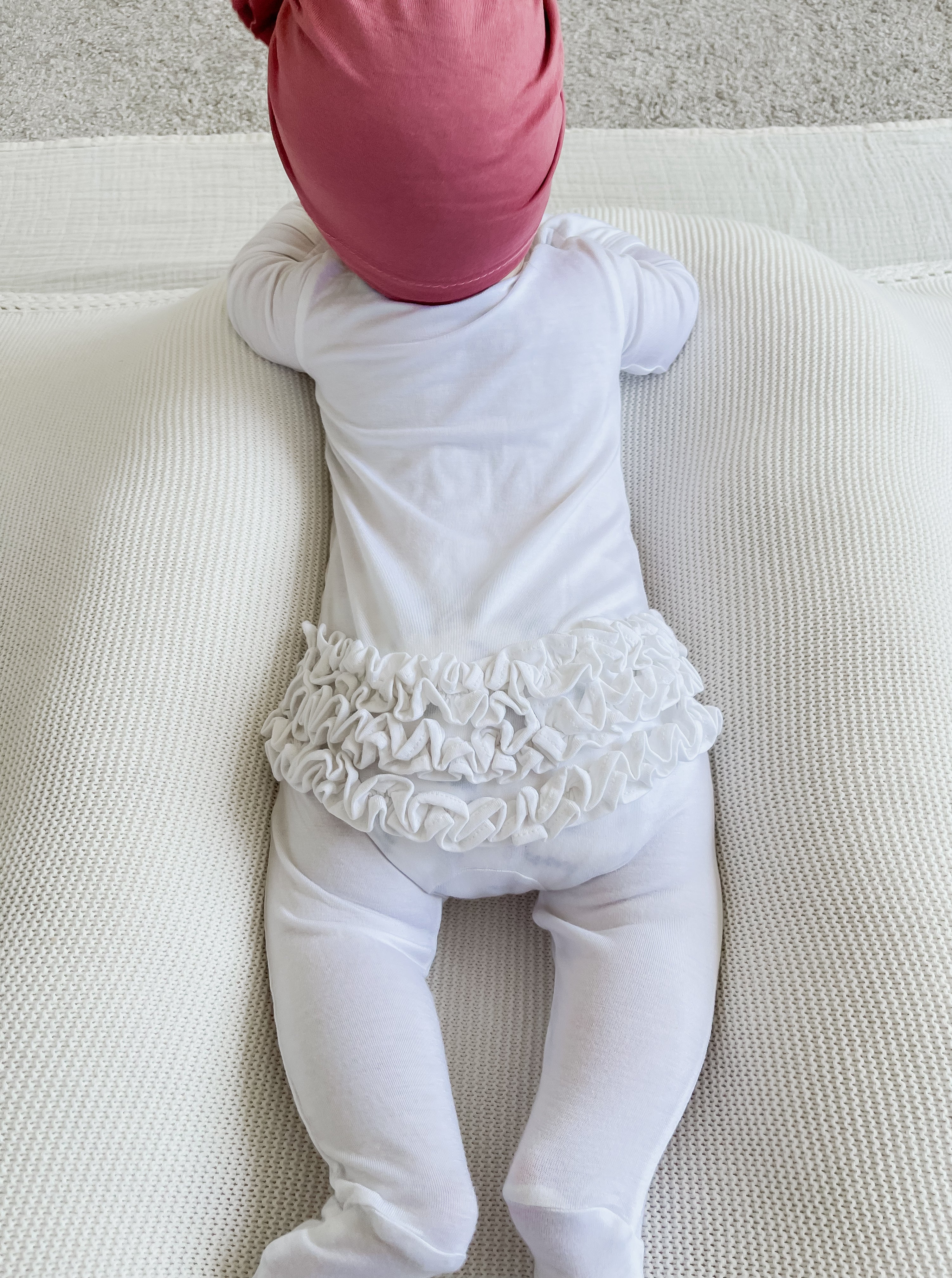 Baby in a white outfit with ruffles, lying on a textured surface, wearing a pink hat.