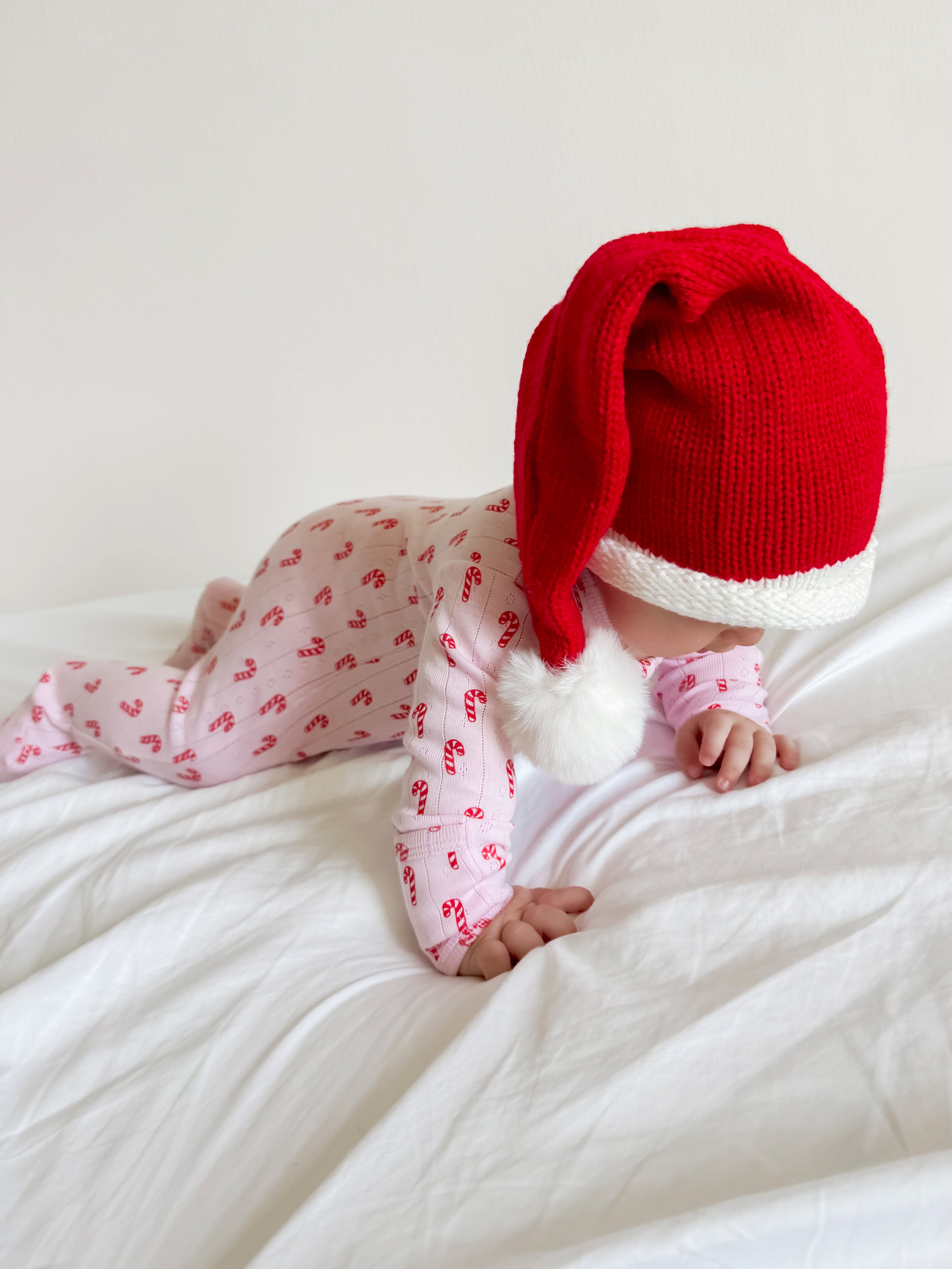 Baby in a red Santa hat crawls on a white blanket, wearing pink pajamas with candy cane patterns.