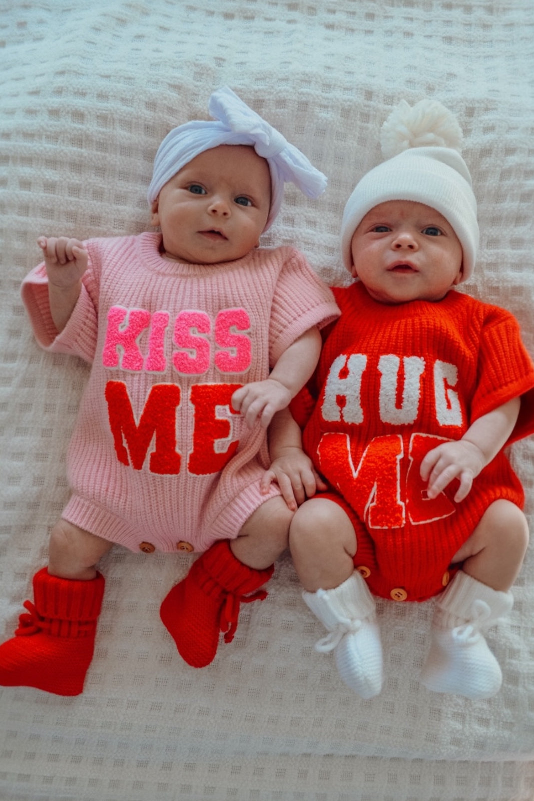 Two infants in colorful knitted outfits that say "KISS ME" and "HUG ME," resting on a soft white blanket.