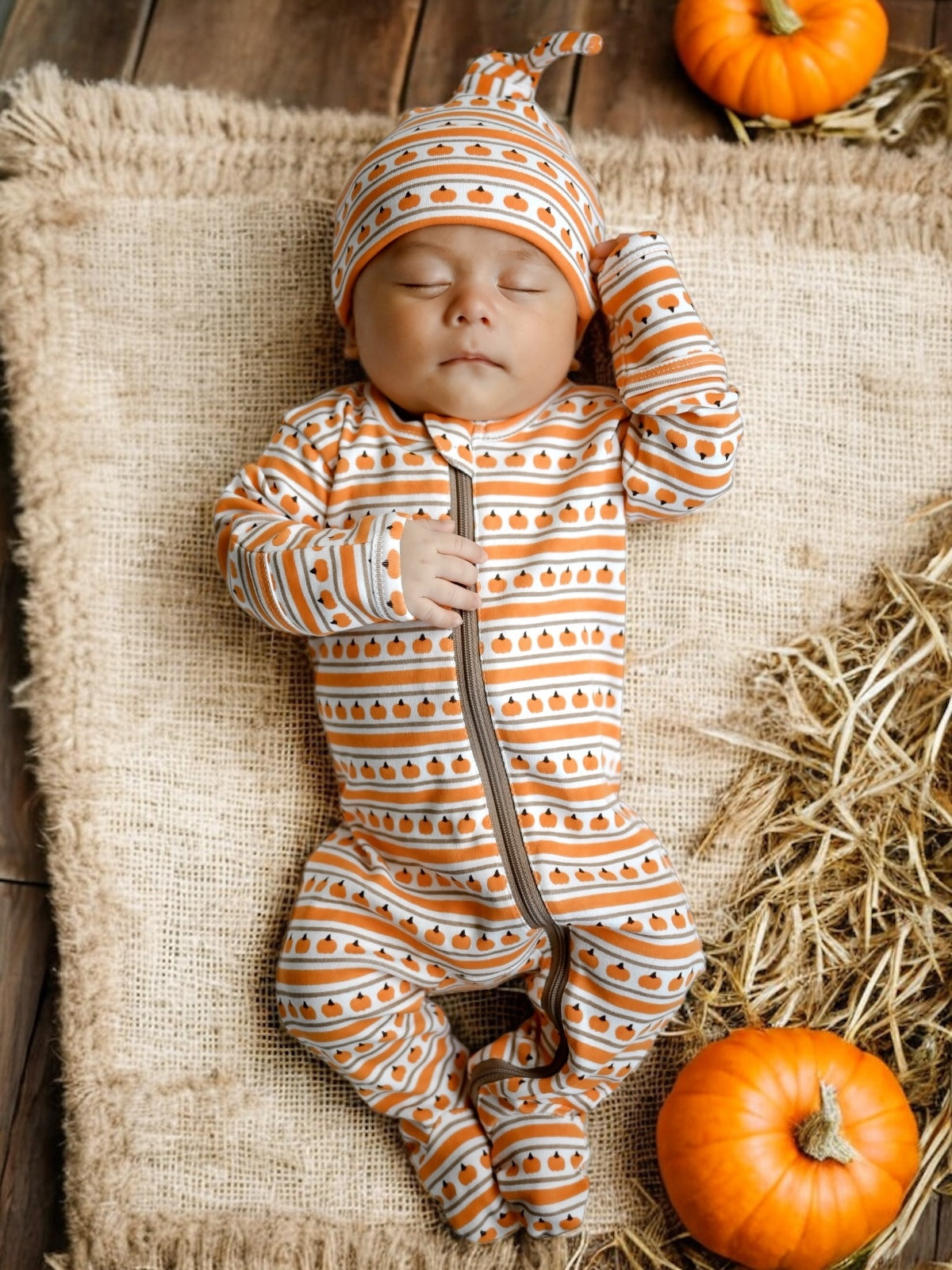 Baby sleeping on a burlap surface, wearing pumpkin-patterned pajamas and hat, with pumpkins nearby.