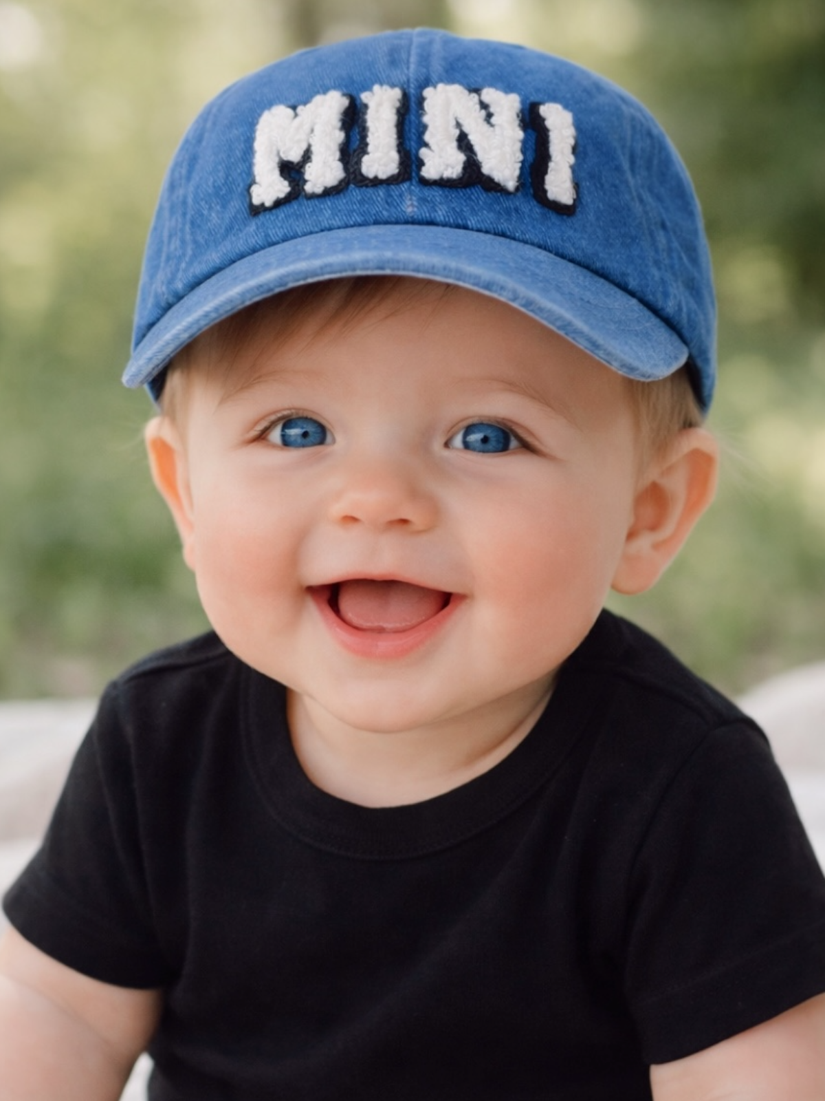 Smiling baby wearing a blue cap with "MINI" on it, sitting outdoors, with soft green background.