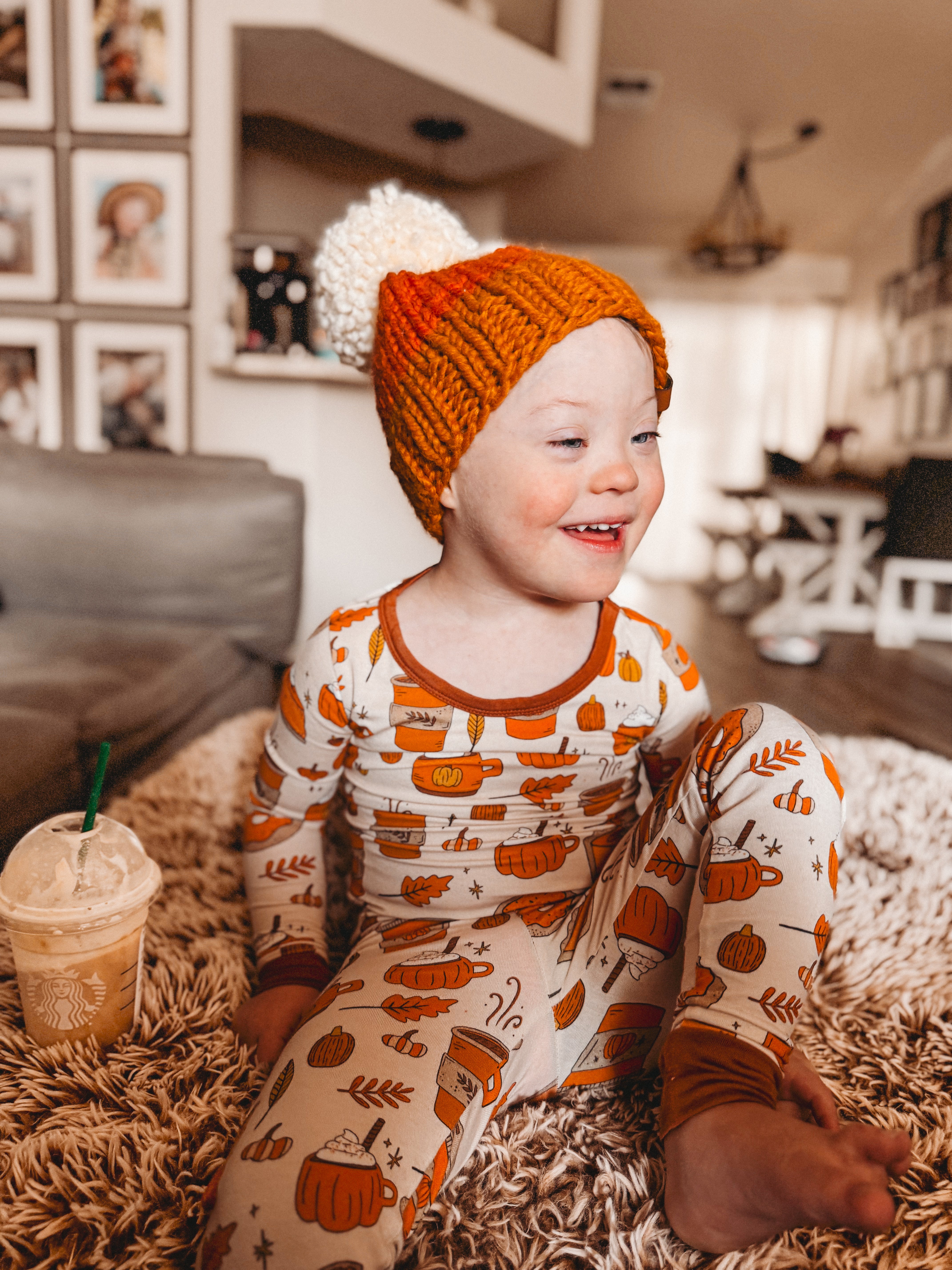 Smiling child in cozy pumpkin-patterned pajamas, wearing a knitted orange hat with a pom-pom, seated on a rug.