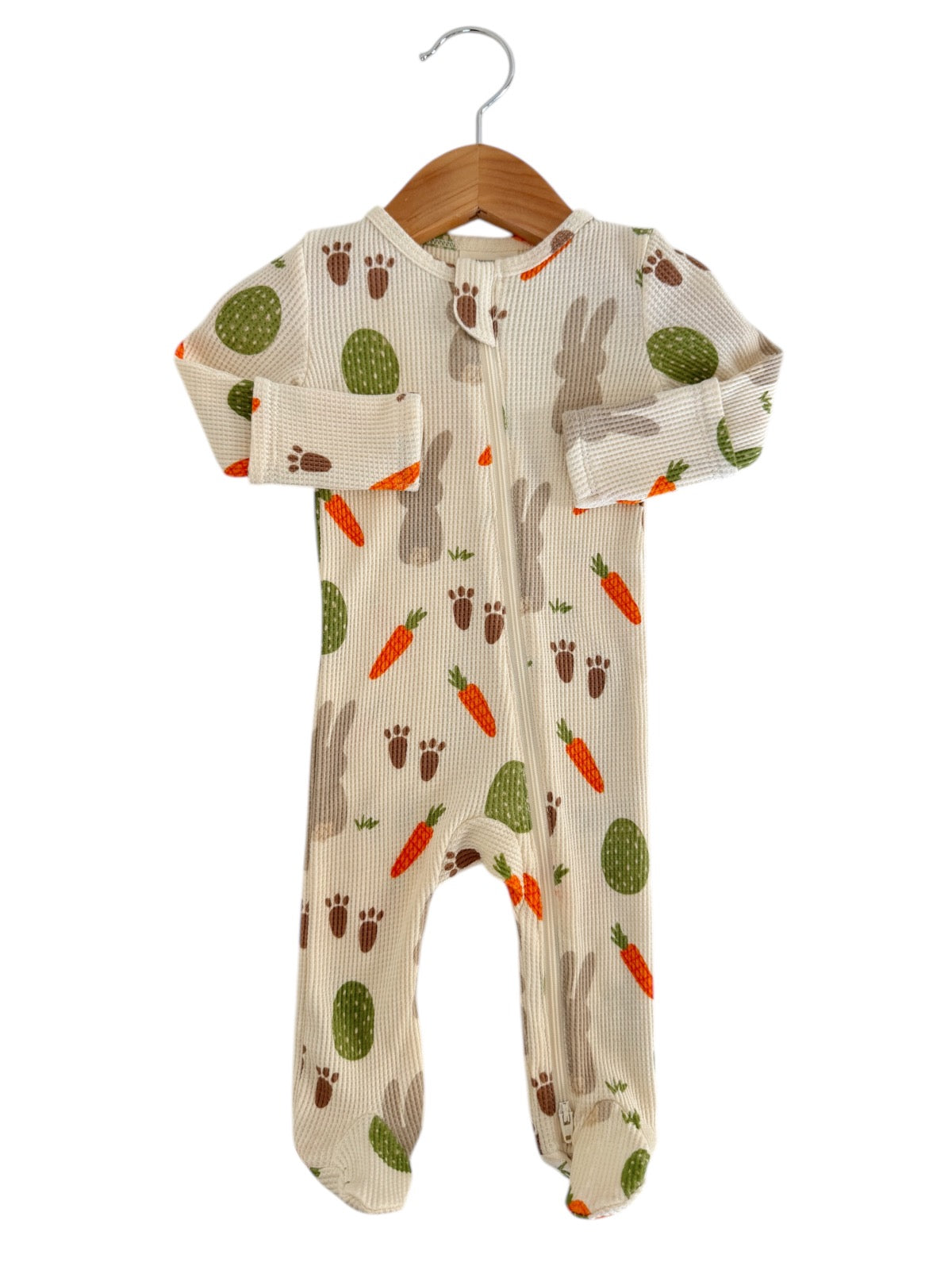 Beige baby onesie with bunny, carrot, and cactus patterns, hanging on a wooden hanger.