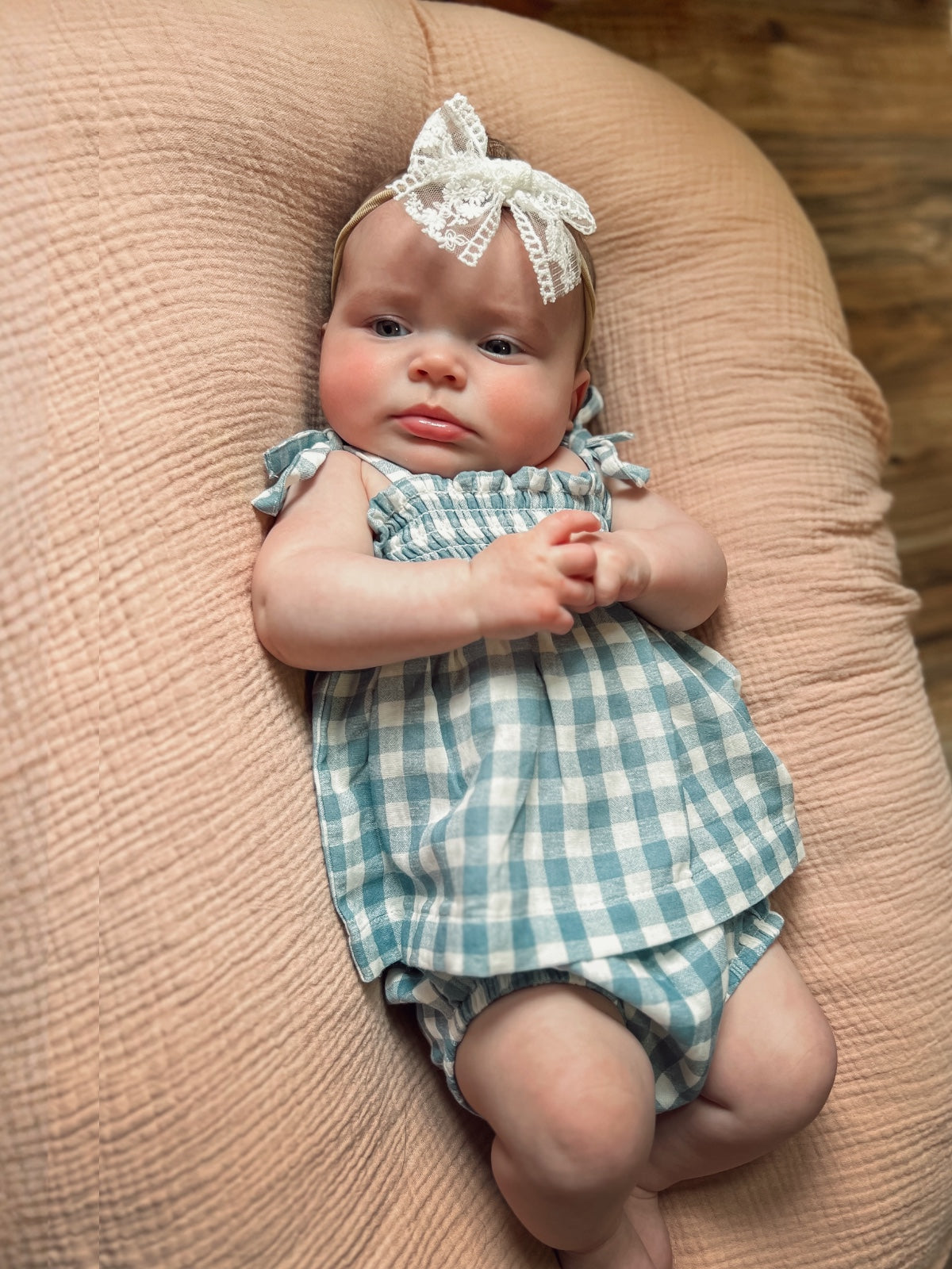 Baby girl in a blue and white checked outfit with a lace headband, resting on a soft, textured surface.