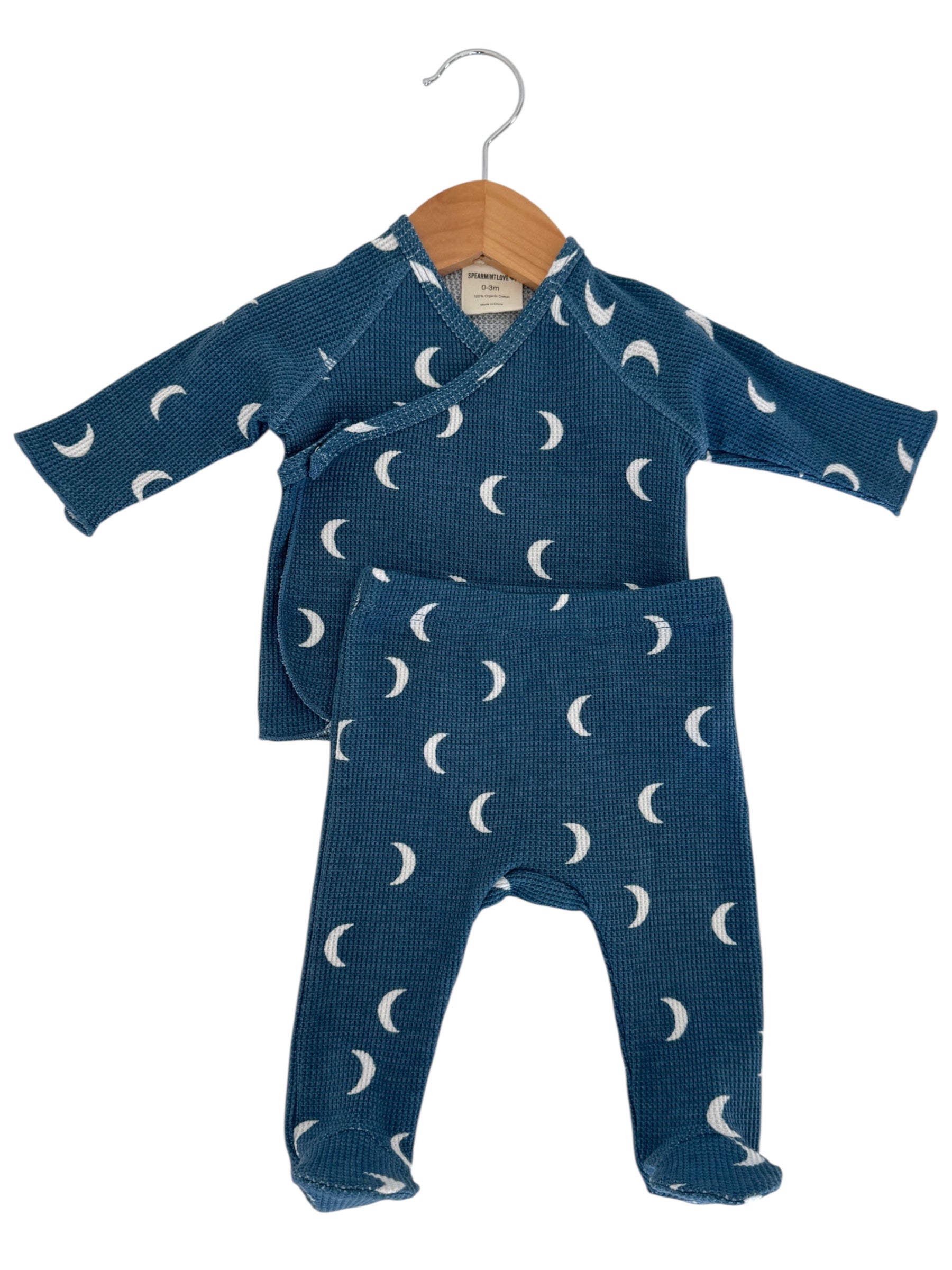 Blue baby outfit with crescent moon pattern, consisting of a wrap top and matching pants hanging on a wooden hanger.