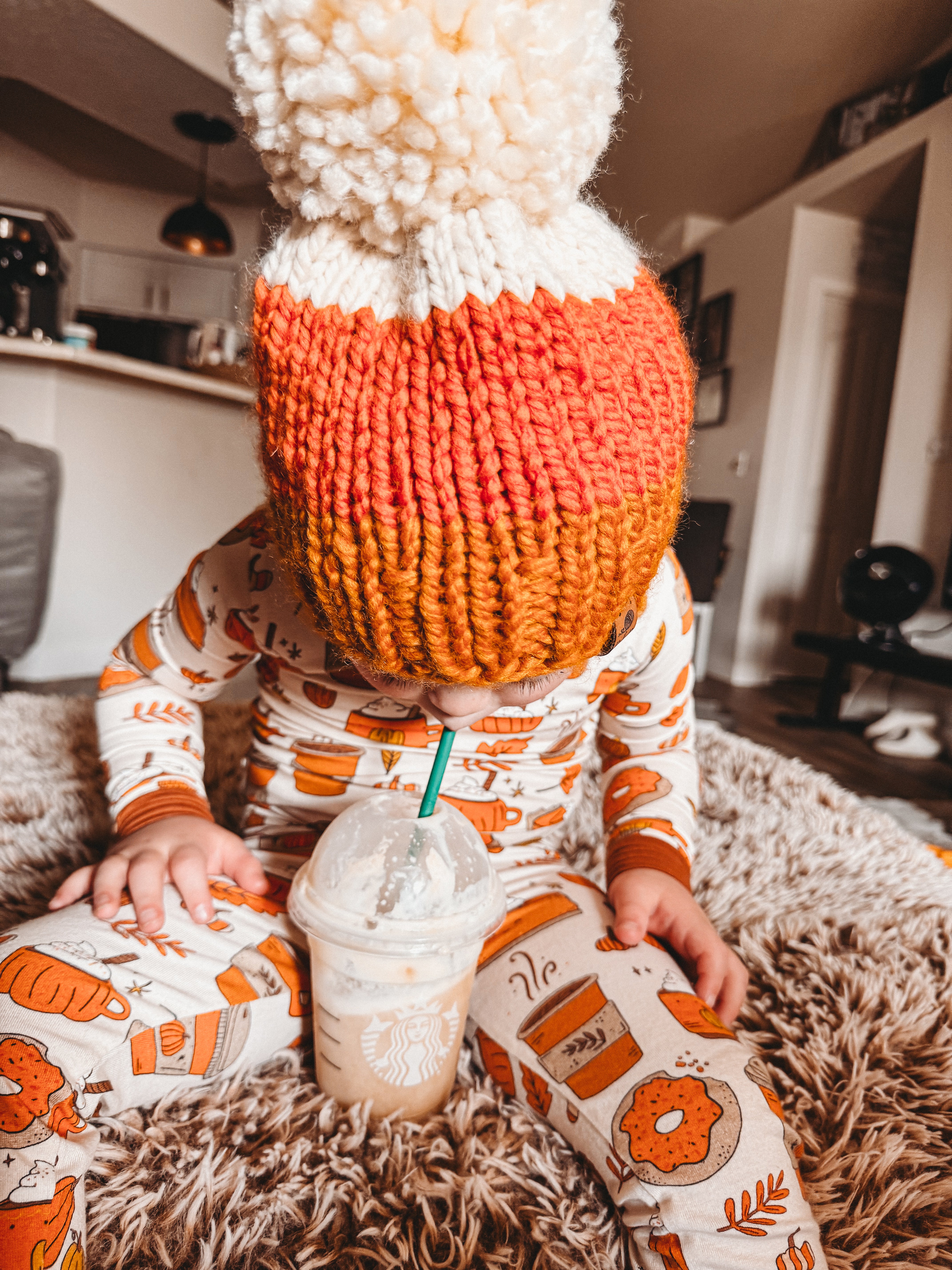 Child wearing a colorful knitted hat sits on a rug, sipping a coffee drink from a Starbucks cup.