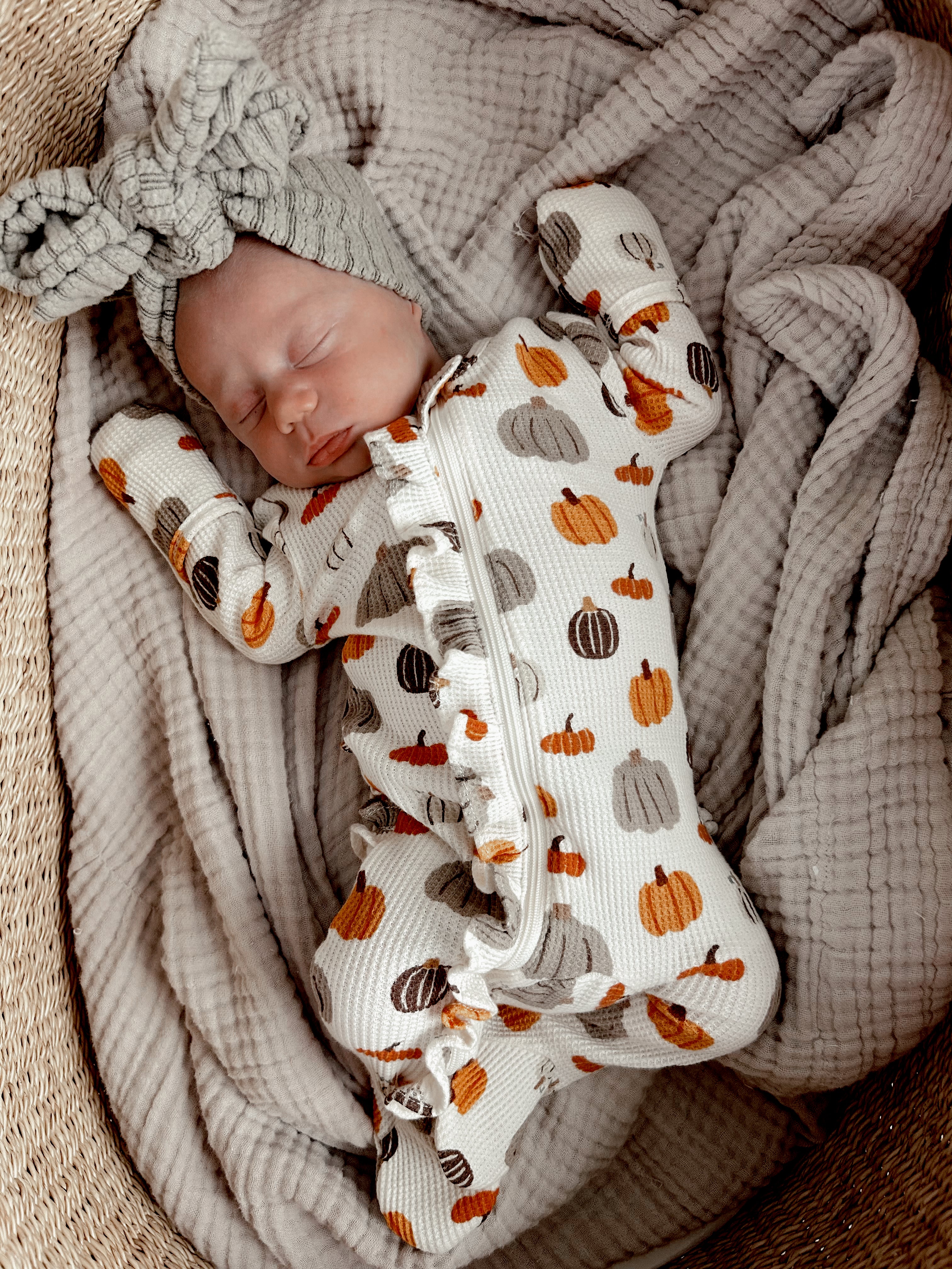Sleeping baby in a pumpkin-patterned outfit and bow headband, nestled in a cozy blanket.