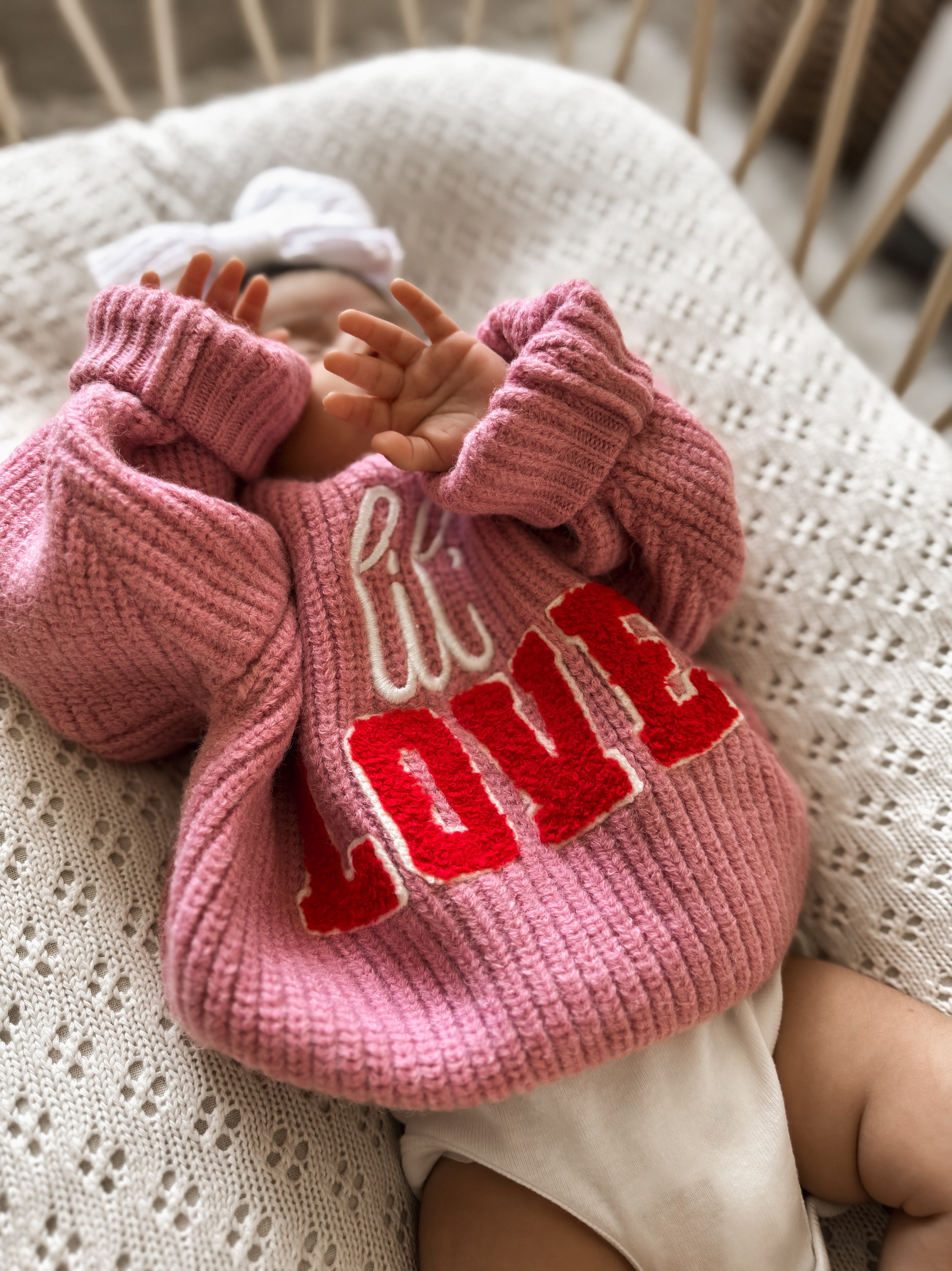 Infant wearing a cozy pink sweater with "LOVE" text, resting on a knitted blanket.