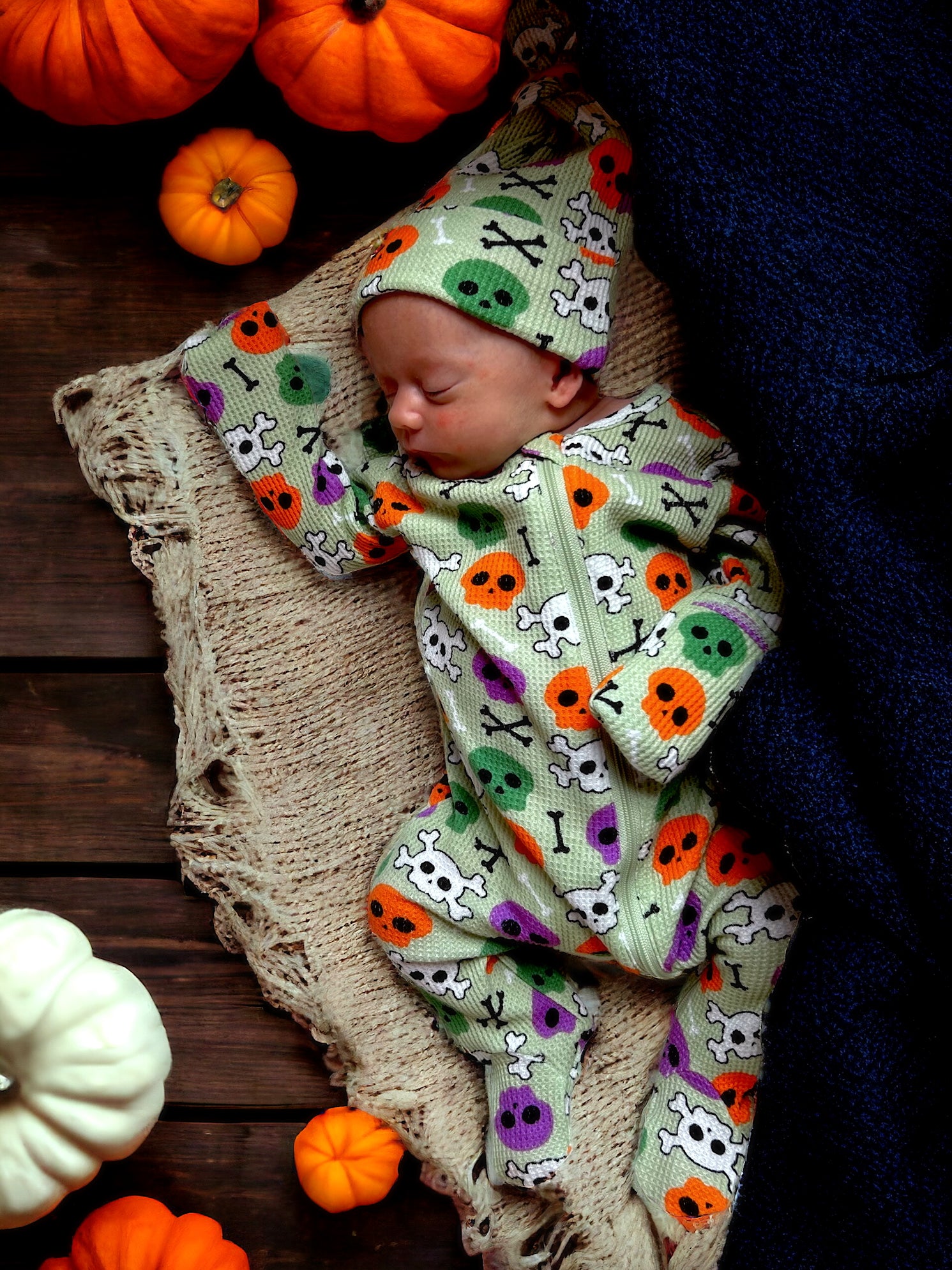 Sleeping baby in a colorful skull-patterned outfit surrounded by pumpkins on a cozy blanket.