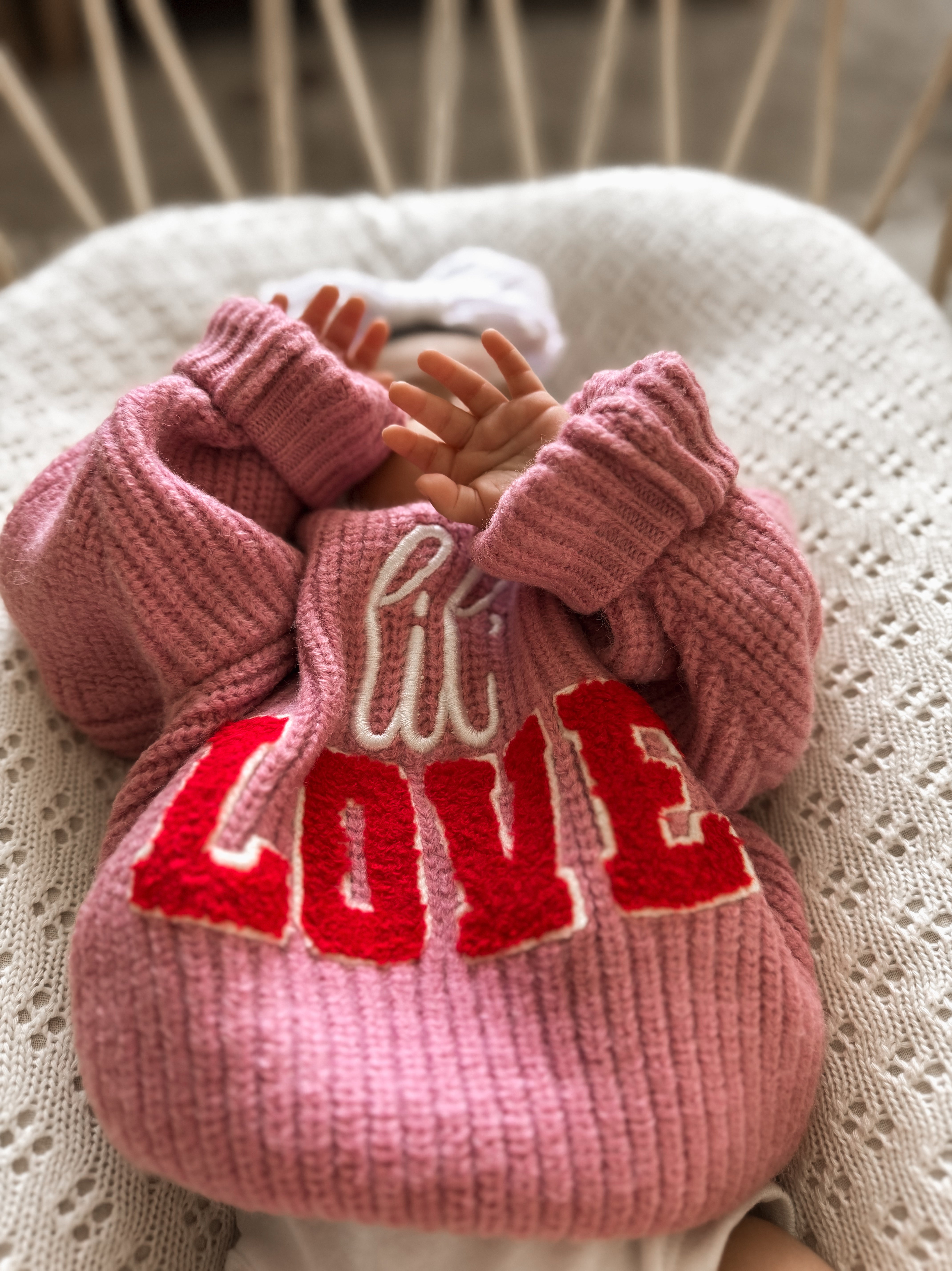 A cozy baby in a pink sweater with "LOVE" text, resting on a knit blanket. Hands are playfully covering their face.