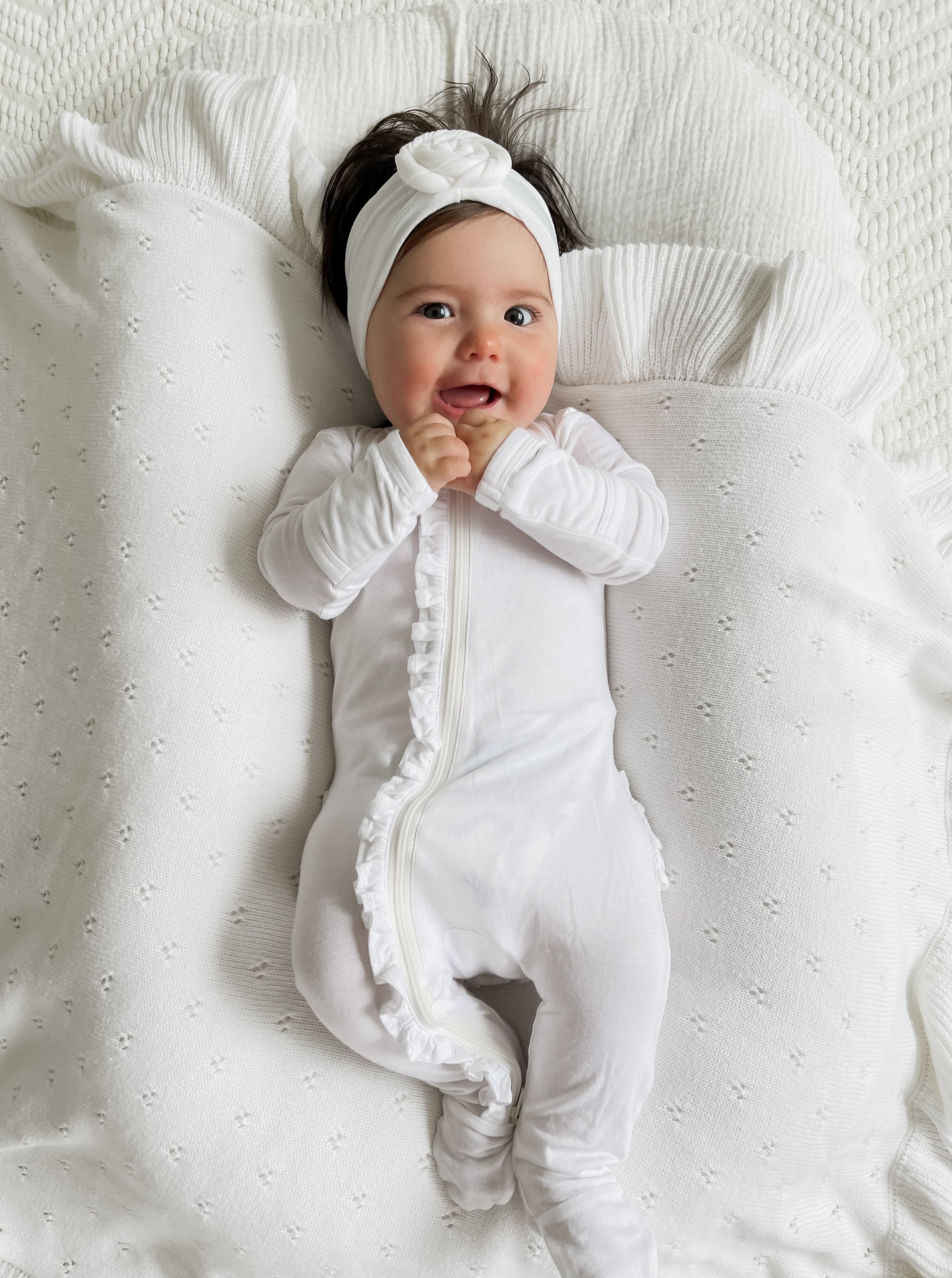 Smiling baby in a white outfit and headband, lying on a textured white blanket with ruffles.