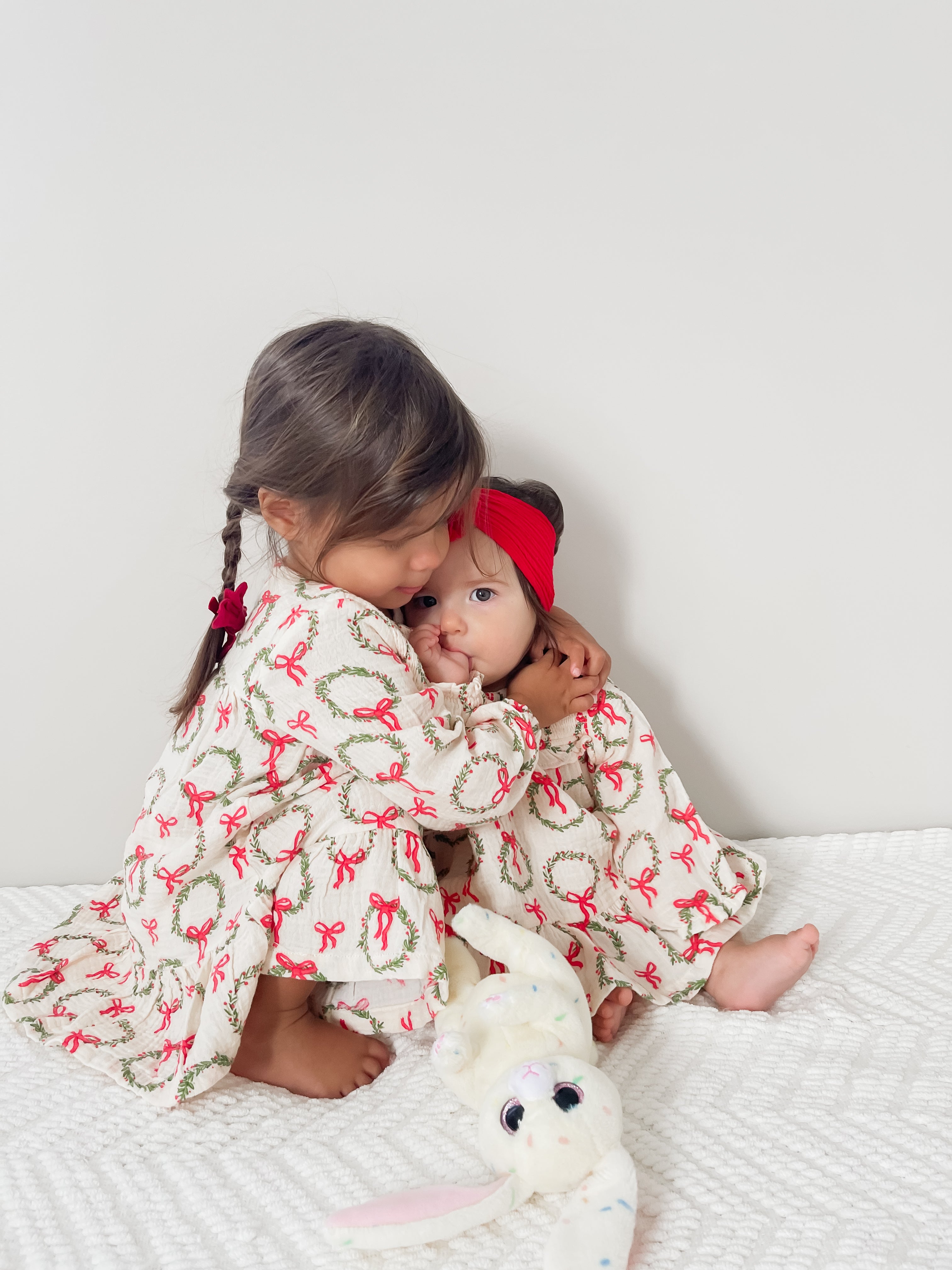 Two young girls in matching dresses embrace, with a plush bunny toy on a textured blanket. Bright, playful scene.