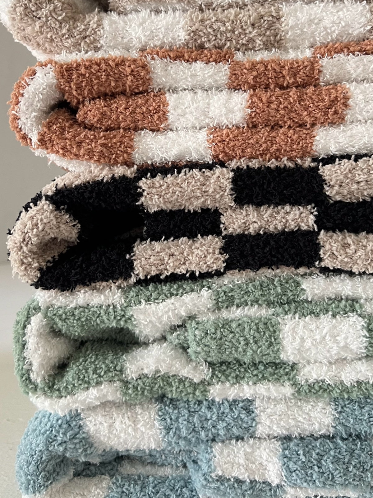Stack of colorful, fluffy blankets in various checkered patterns and hues of green, brown, black, and cream.