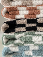 Stack of colorful, fluffy blankets in various checkered patterns and hues of green, brown, black, and cream.