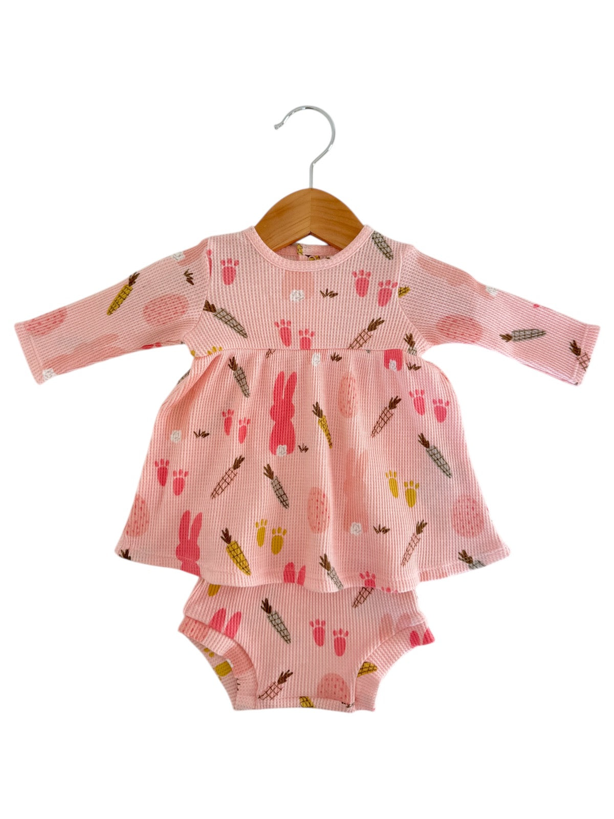 Pink baby dress with bunny and fruit patterns, paired with matching bloomer shorts, hanging on a wooden hanger.