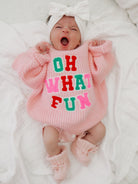 Baby in pink sweater with colorful letters "OH WHAT FUN," yawning, wearing a bow headband and soft socks.