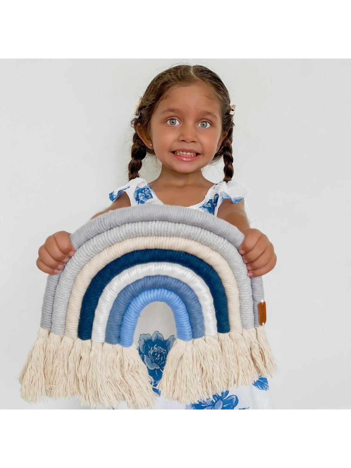 Smiling young girl holding a colorful fabric rainbow decoration with fringes, wearing a floral dress.