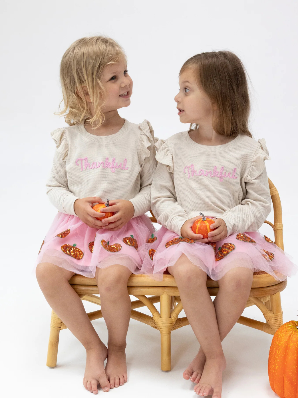Two smiling girls in matching outfits with "Thankful" text, holding small pumpkins, sitting on a chair.