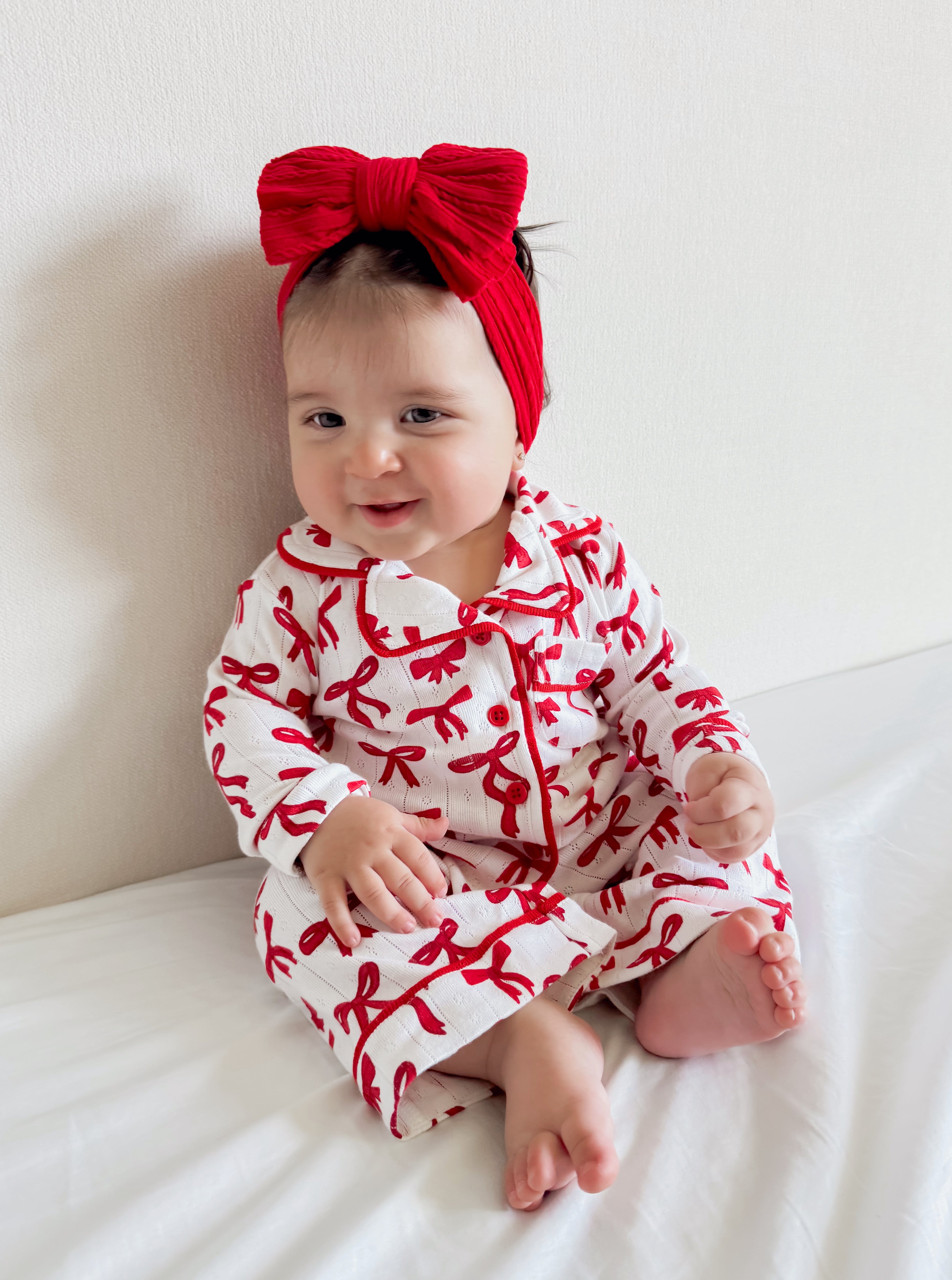 Smiling baby girl in red bow headband and patterned outfit sitting on a bed against a neutral background.