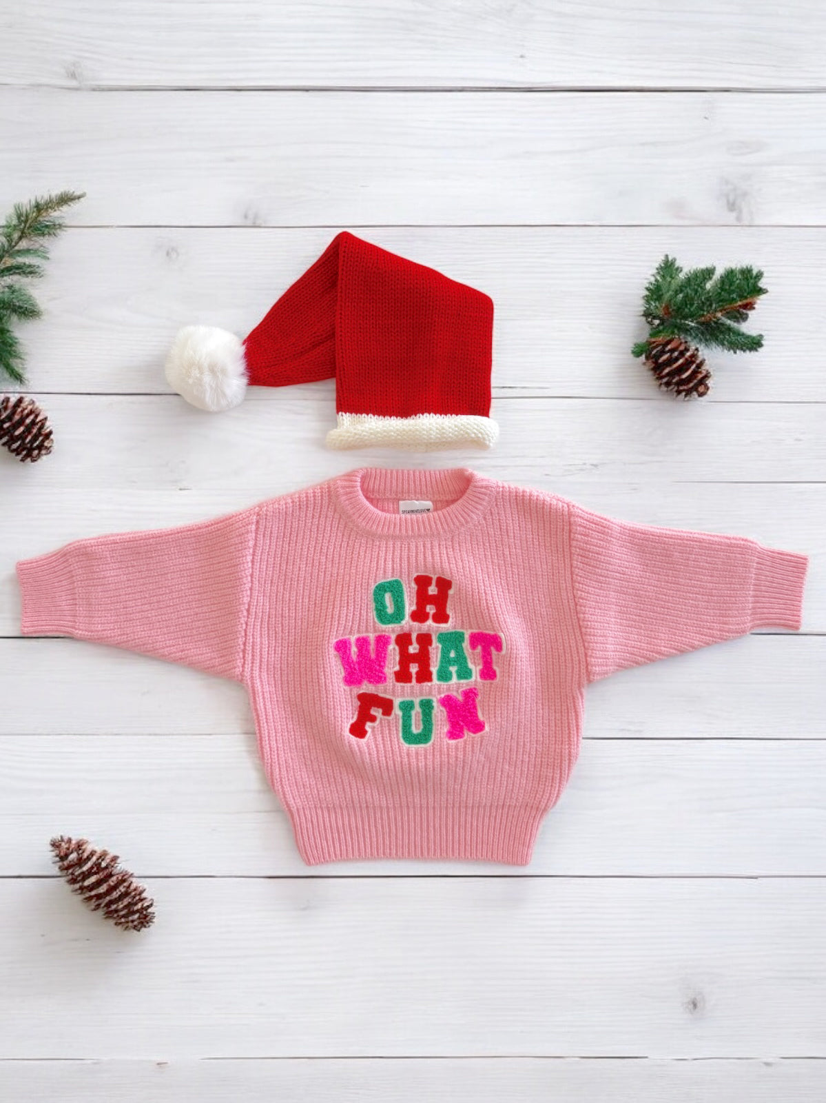 Pink sweater with "OH WHAT FUN" lettering, red Santa hat, pinecones, and greenery on a white wooden background.